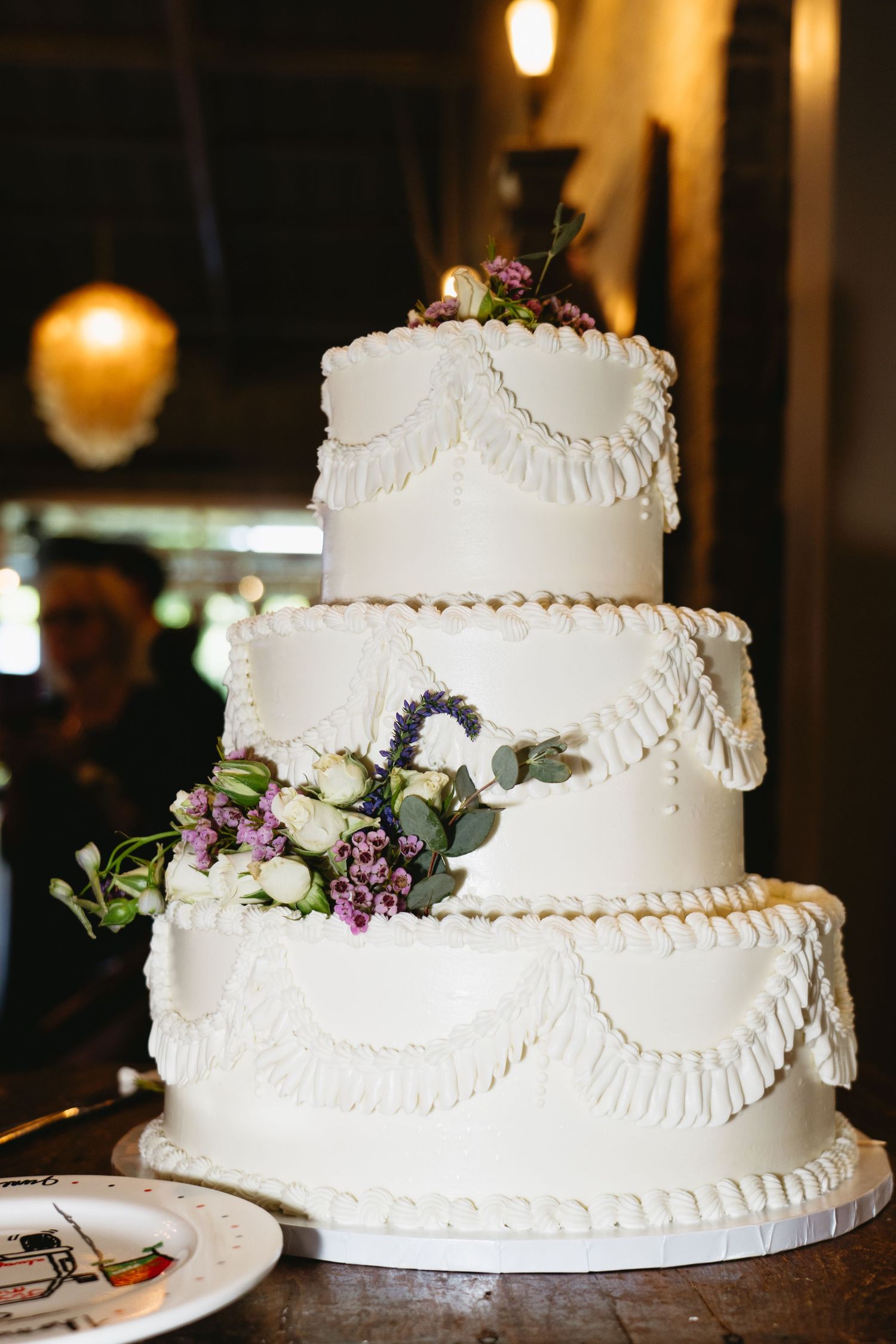 Elegant three-tier white wedding cake with ruffled frosting and purple and white floral decorations on dark background.