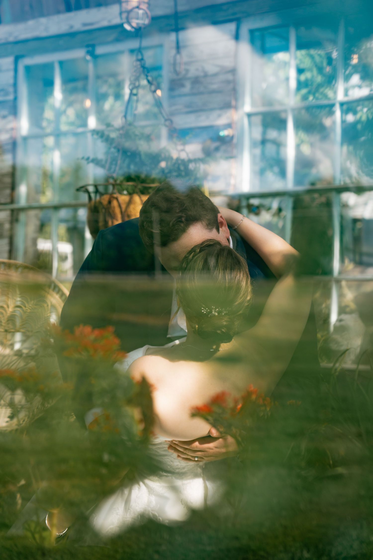Romantic couple sharing a kiss captured through glass window reflection surrounded by greenery.