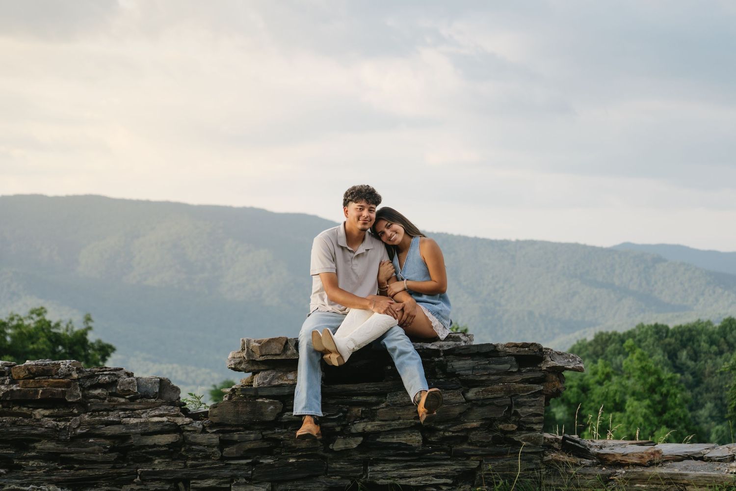 A couple sits closely together on a stone wall with stunning mountain views in the background.