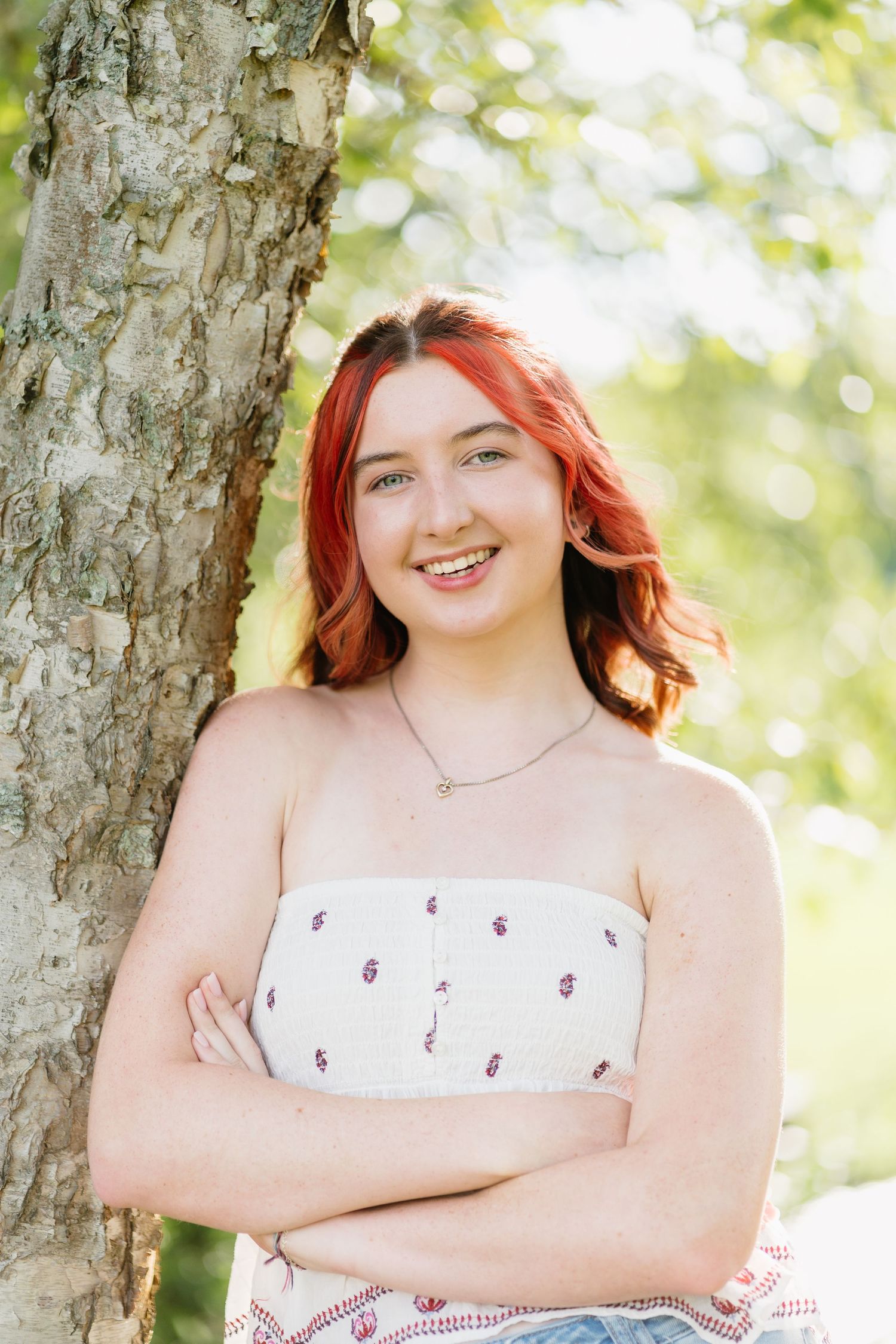 A smiling teenage girl with red hair leans against a tree trunk with arms crossed outdoors.