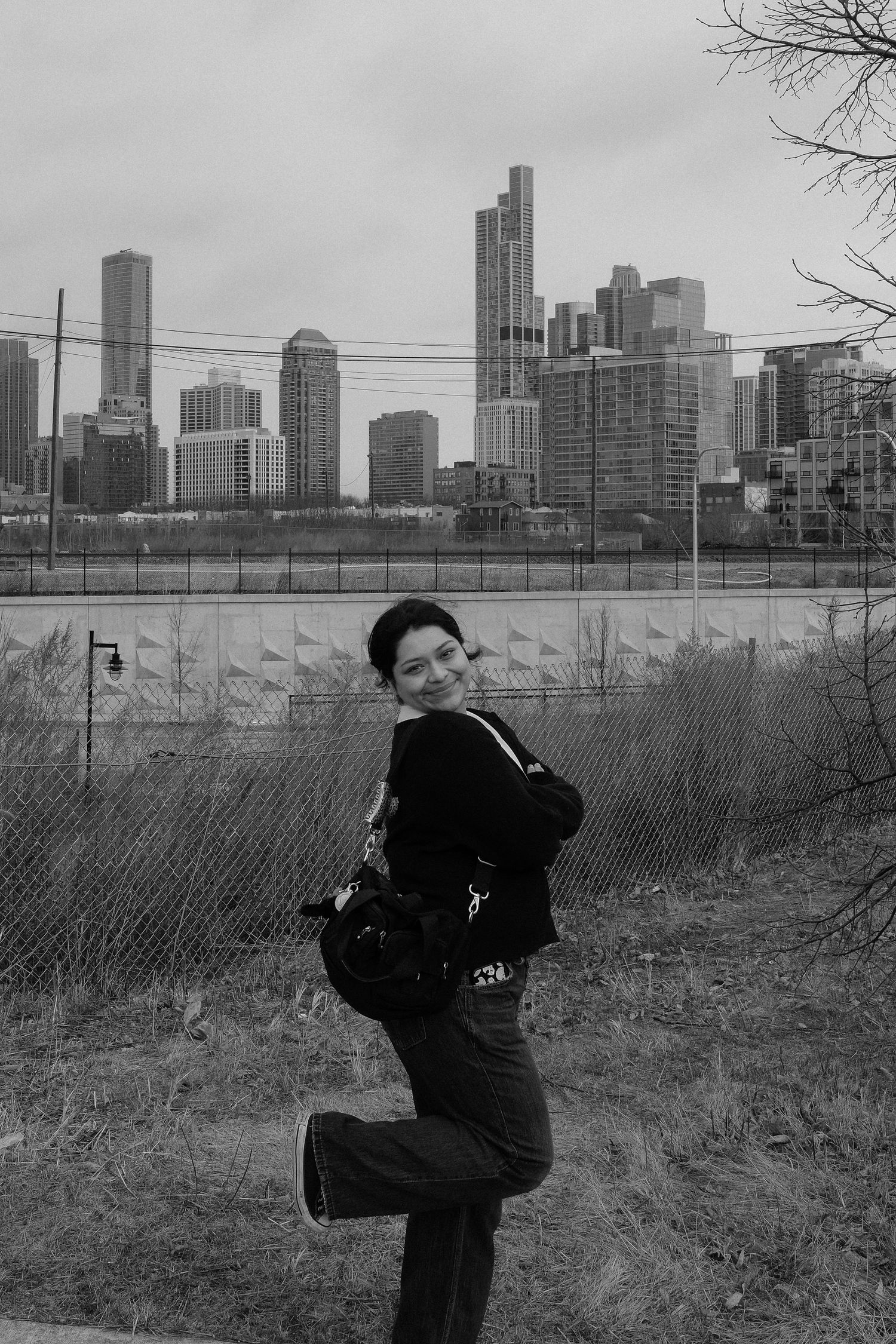 A woman poses playfully in front of a city skyline with skyscrapers visible behind a chain-link fence in black and white.