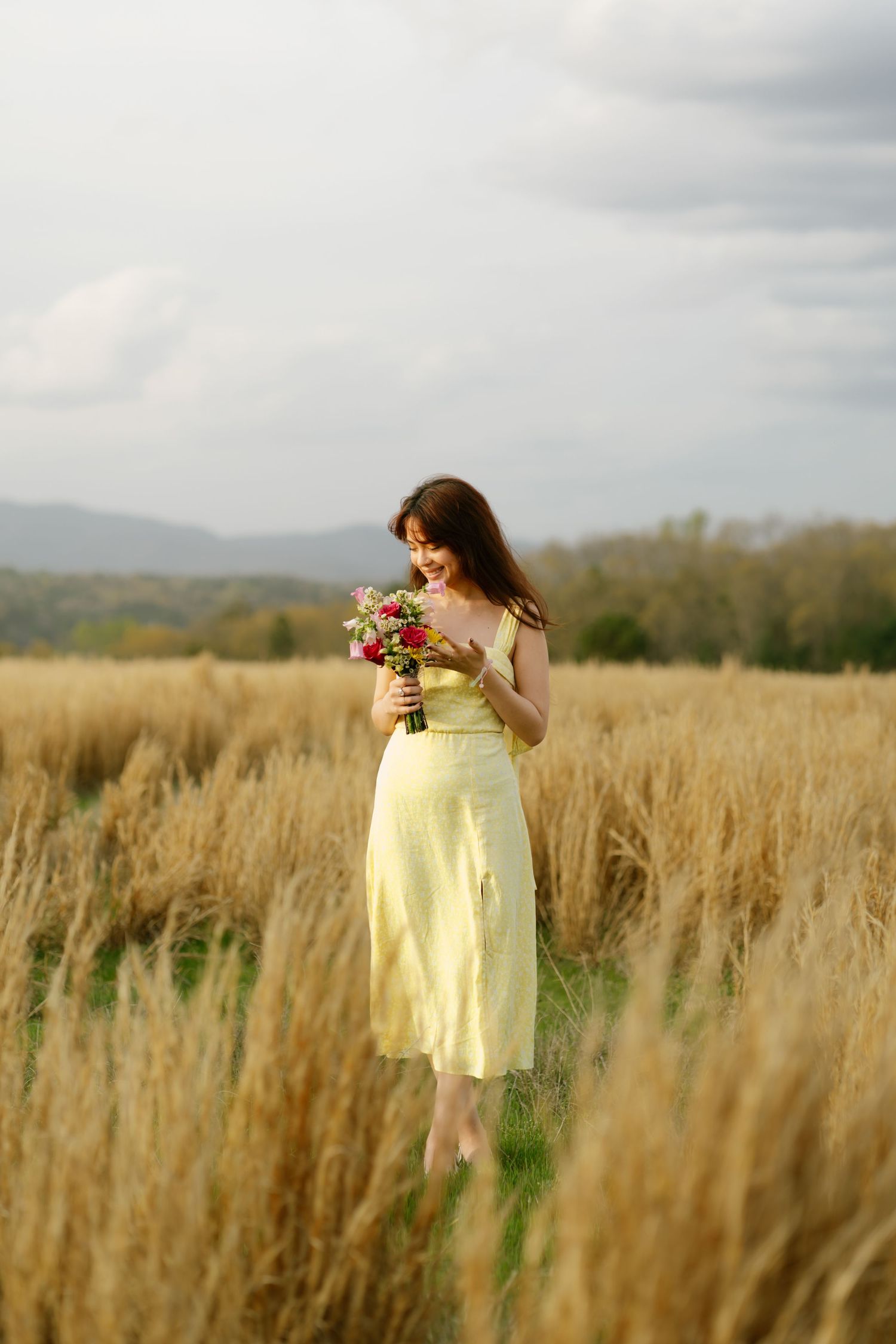 Young woman in yellow sundress smells colorful wildflower bouquet while standing in golden wheat field under cloudy sky.