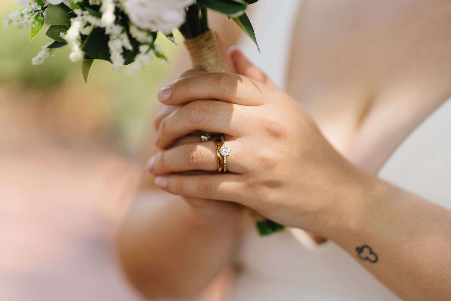 Close-up of bride holding wedding bouquet with gold engagement ring and small heart tattoo on wrist.