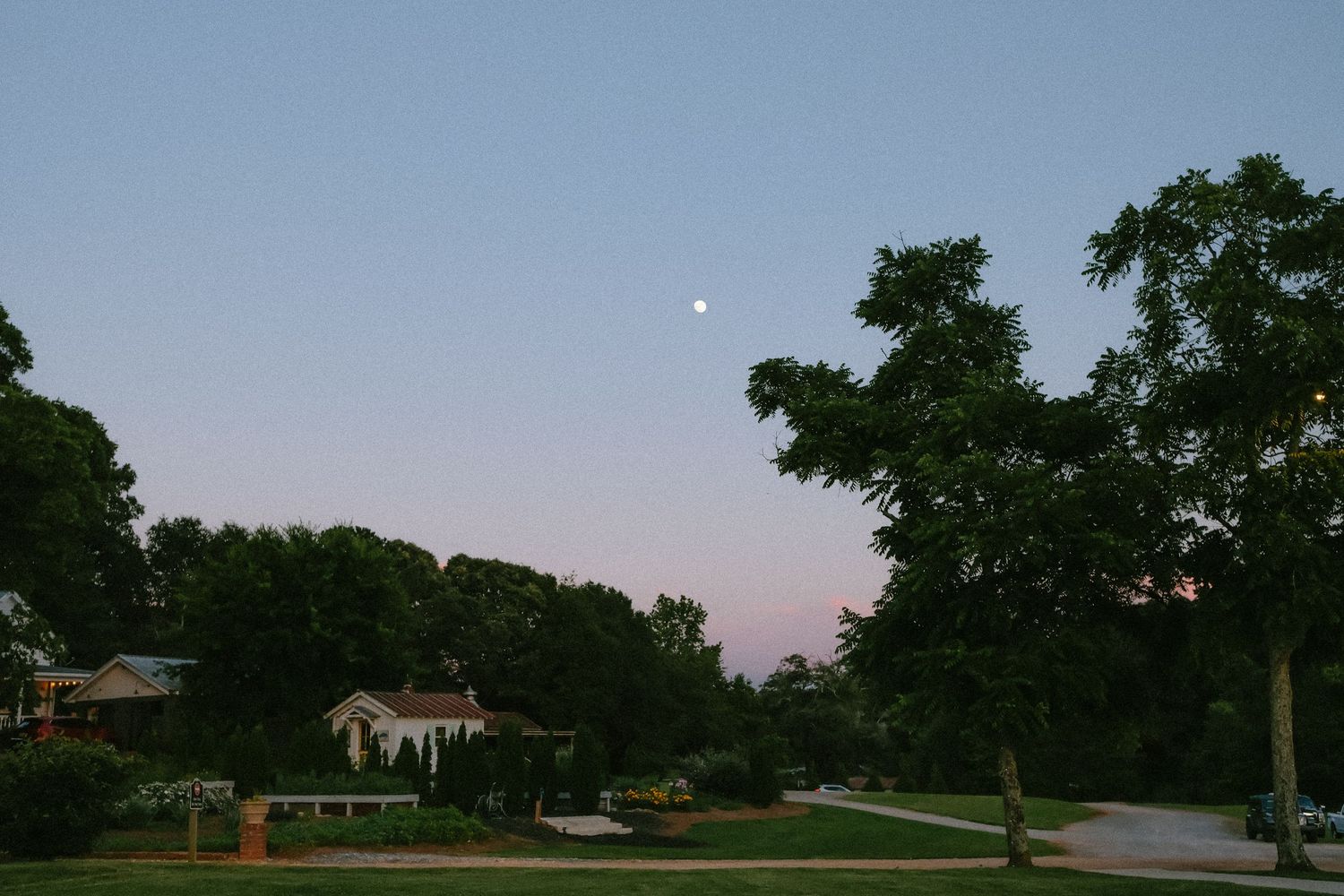 Peaceful dusk landscape with a full moon rising over green trees and a quiet suburban neighborhood.