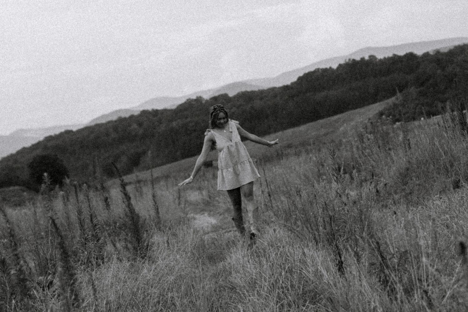 A black and white photo of a woman in a short dress walking through a grassy hillside landscape.
