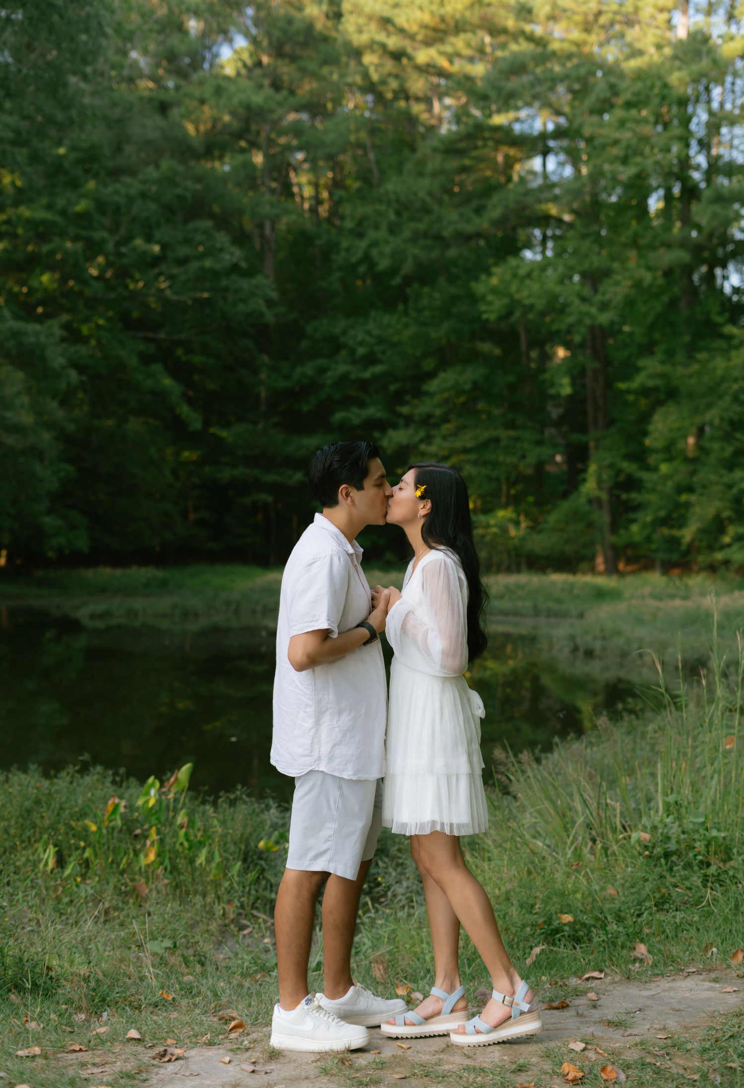 A couple shares a romantic kiss outdoors near a pond surrounded by lush green trees, both dressed in white outfits.
