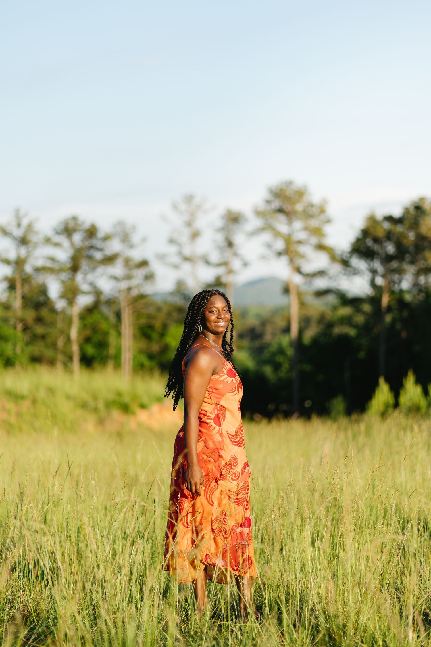 Smiling Black woman with braided hair stands in lush green meadow wearing orange floral maxi dress at sunset.