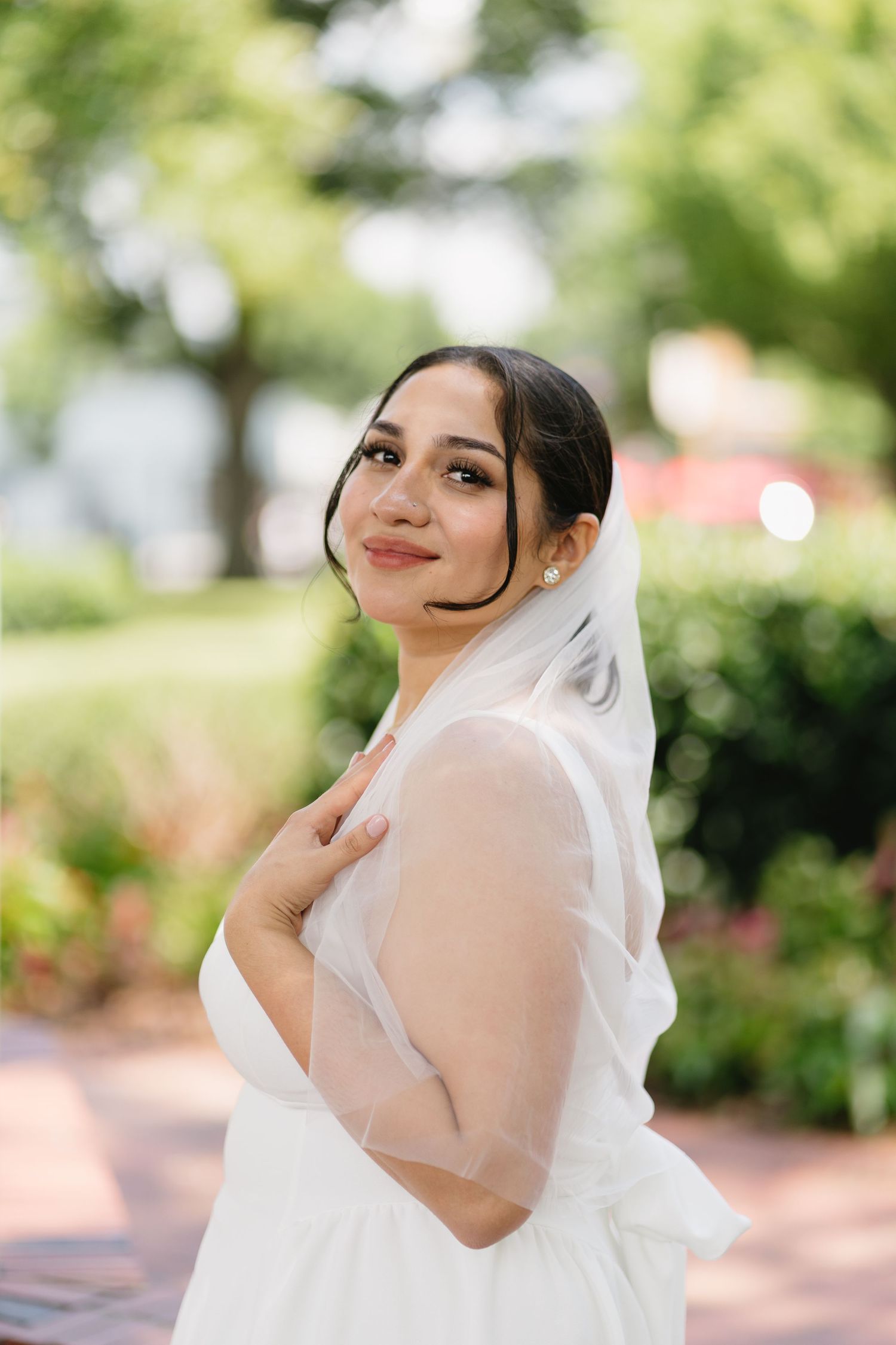 A beautiful bride in a white gown and veil poses gracefully in a sunlit outdoor garden setting.