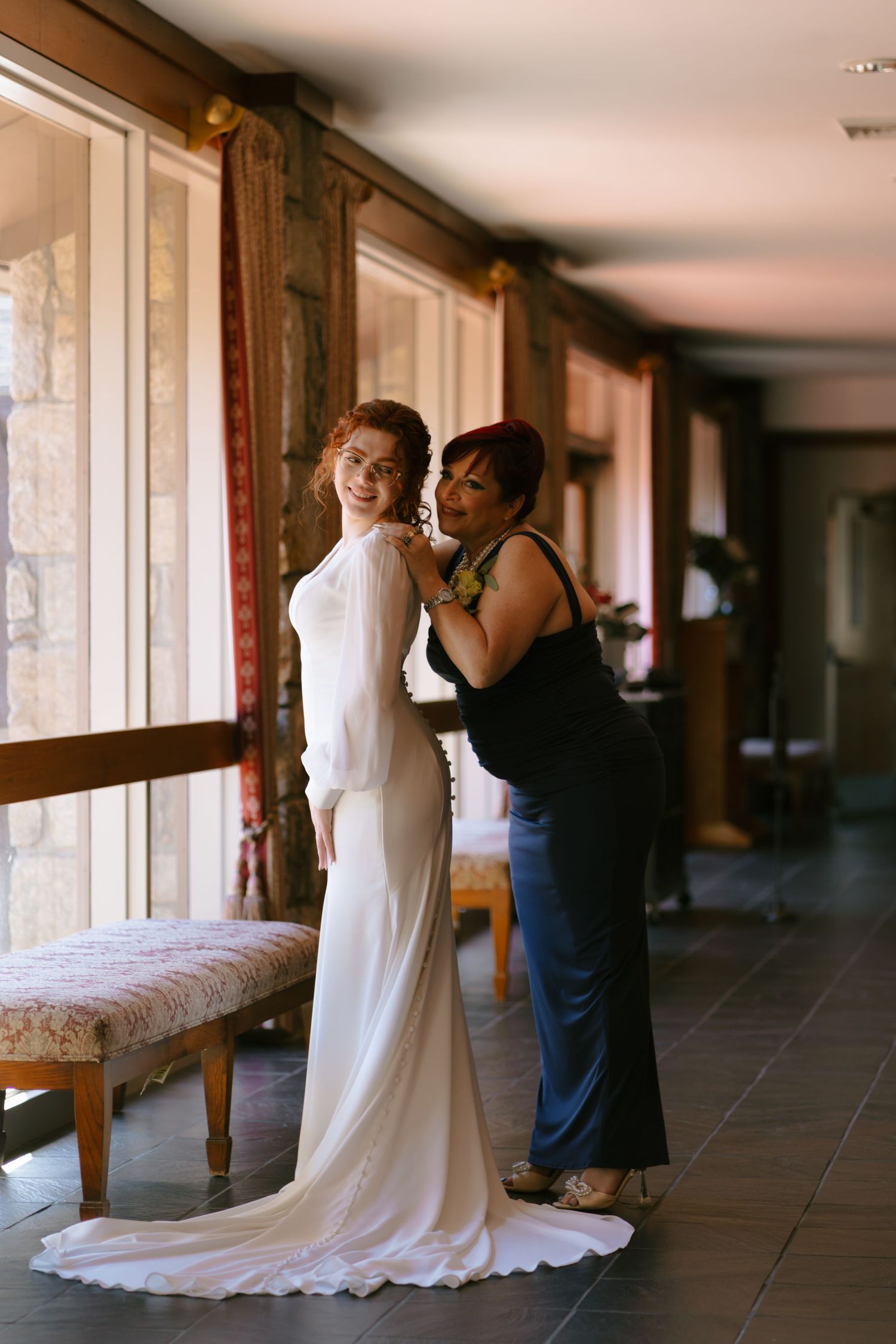 Mother helps fasten the back of brides elegant white gown in warmly lit dressing room.