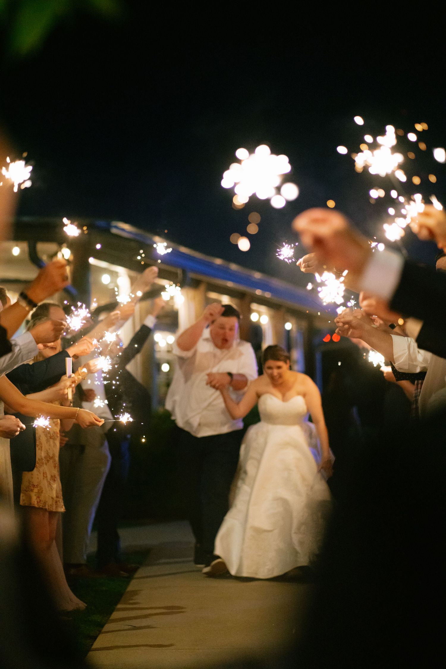 Bride and groom exit through a sparkler tunnel surrounded by cheering guests at a nighttime wedding reception.