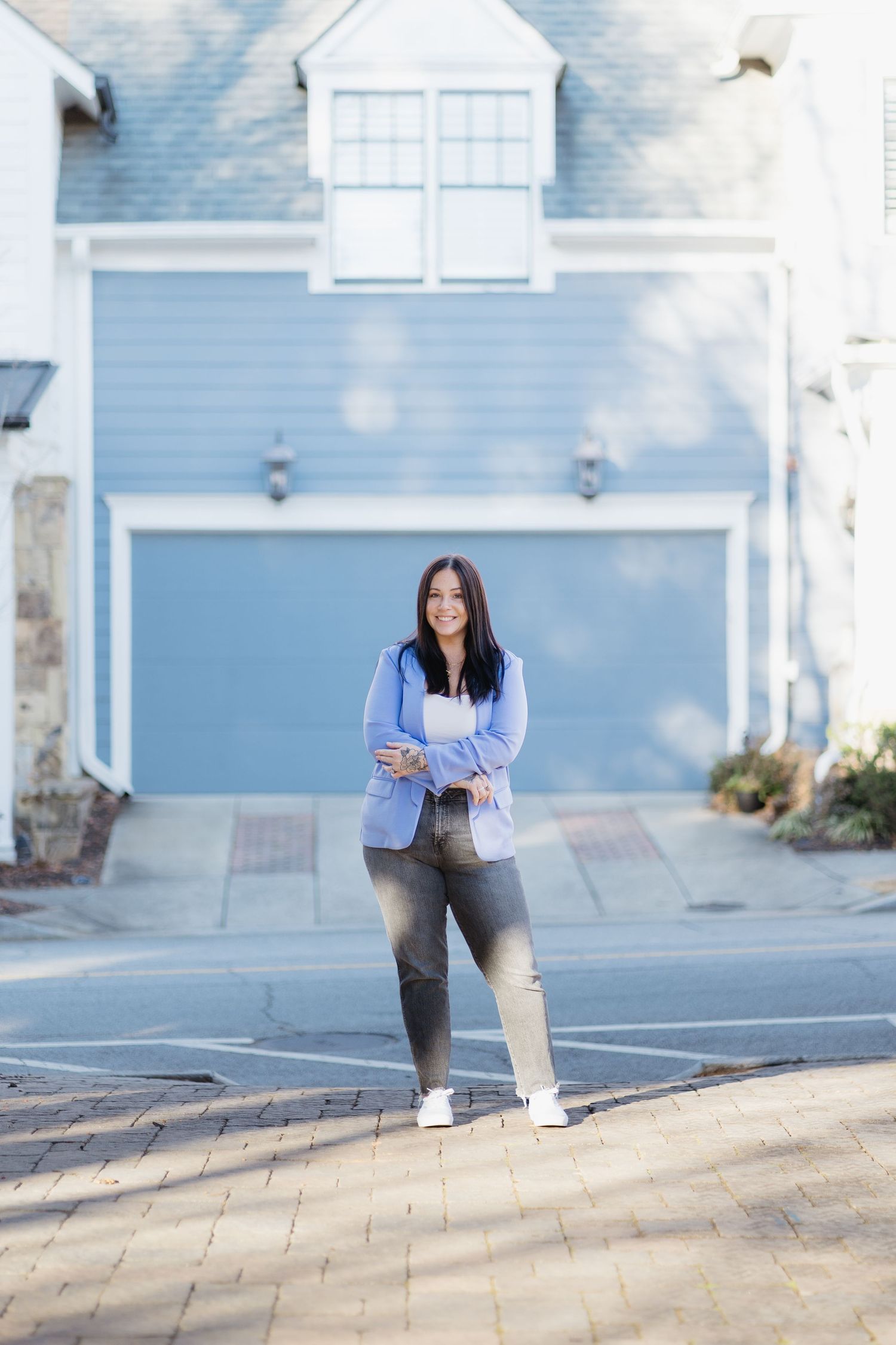 Confident woman in blue blazer and ripped jeans stands arms crossed in front of a blue garage.