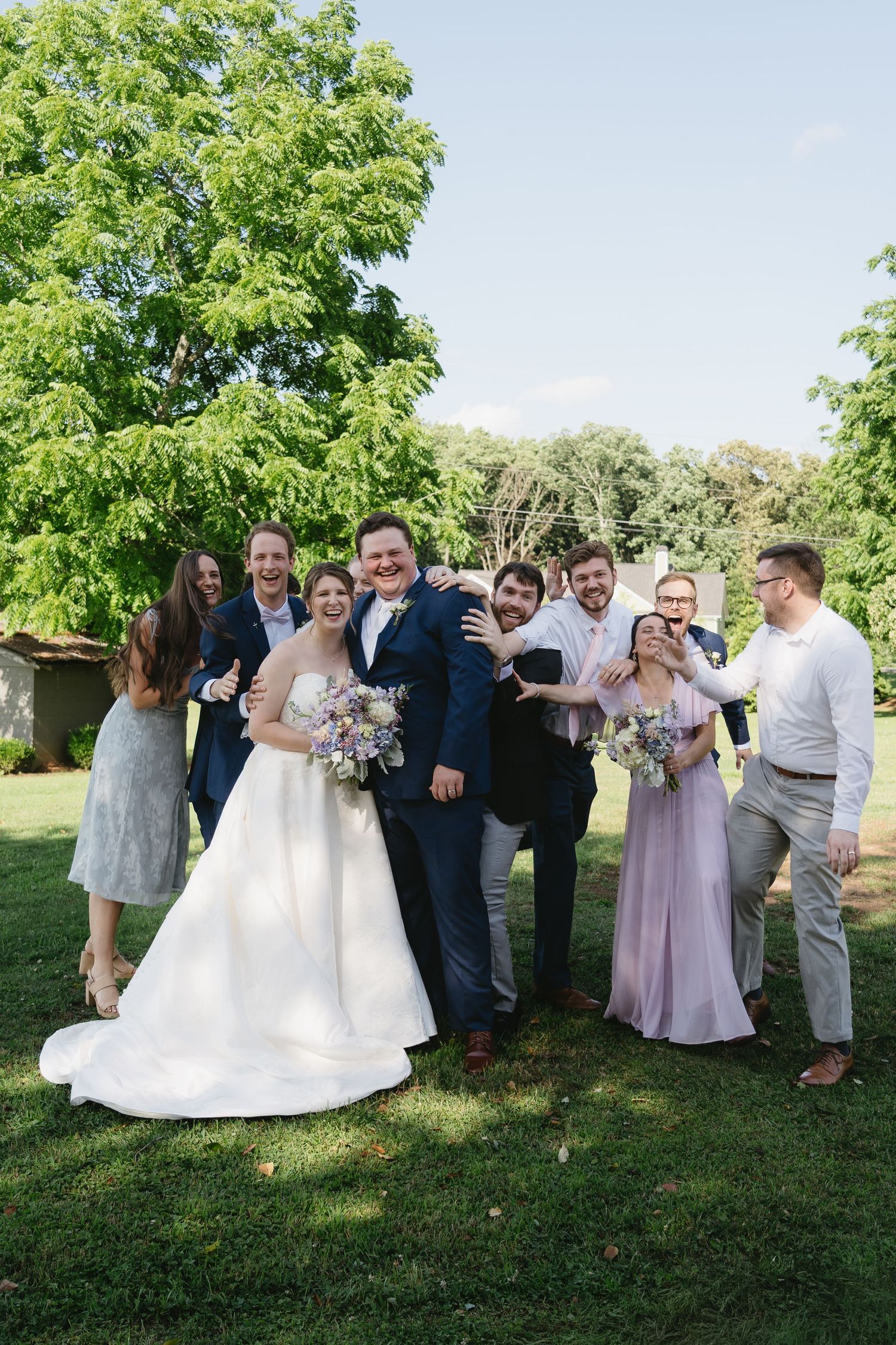 A joyful wedding party poses outdoors on green grass, with the bride in white and groom in navy surrounded by smiling guests.