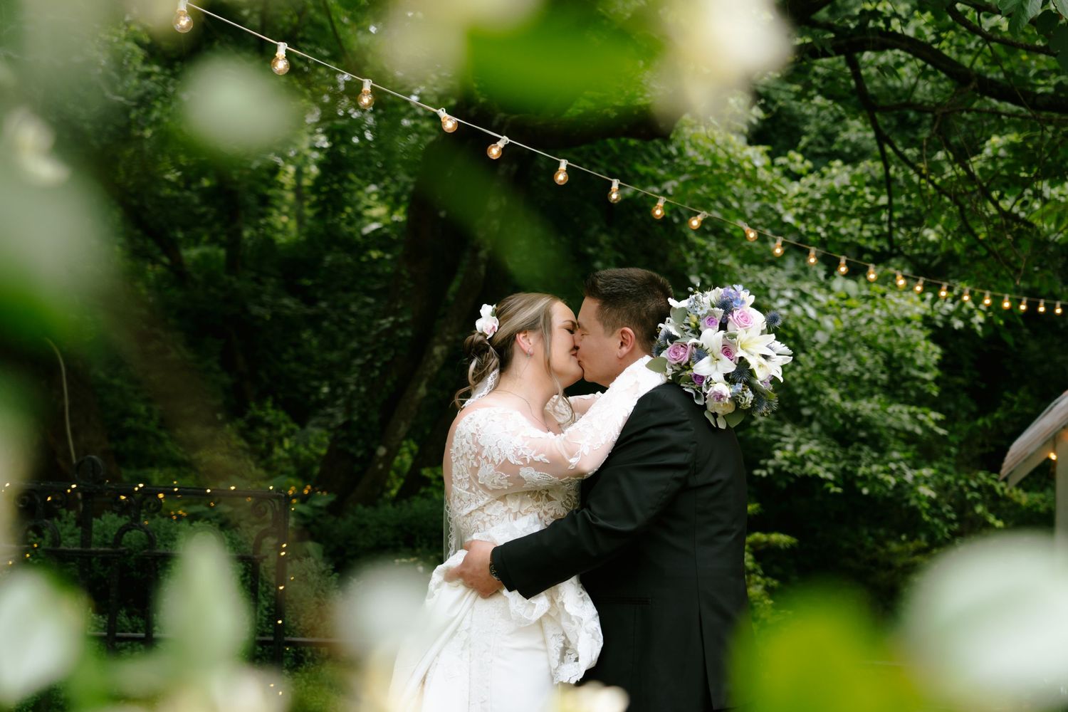 Bride and groom share a romantic kiss outdoors, surrounded by lush greenery and string lights at their garden wedding.