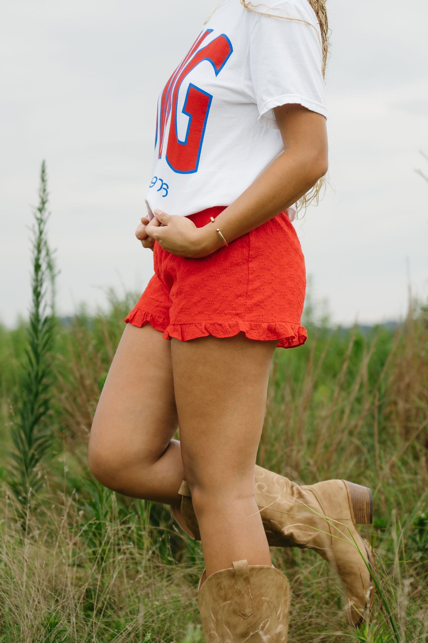A woman in red ruffled shorts, a graphic tee, and tan cowboy boots stands in a green field.