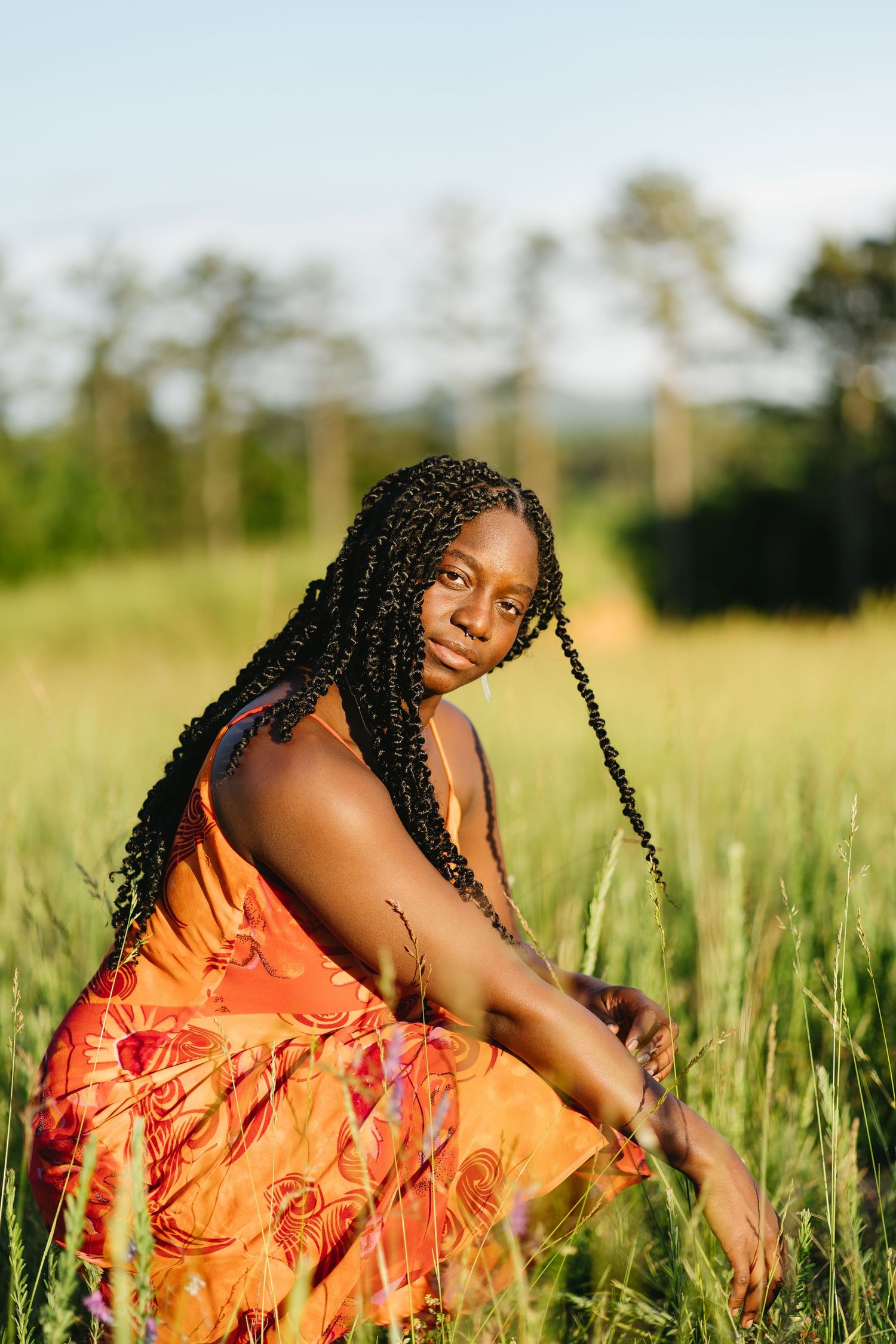 Young Black woman with long braided hair sits in green field wearing vibrant orange floral sundress in golden sunlight.