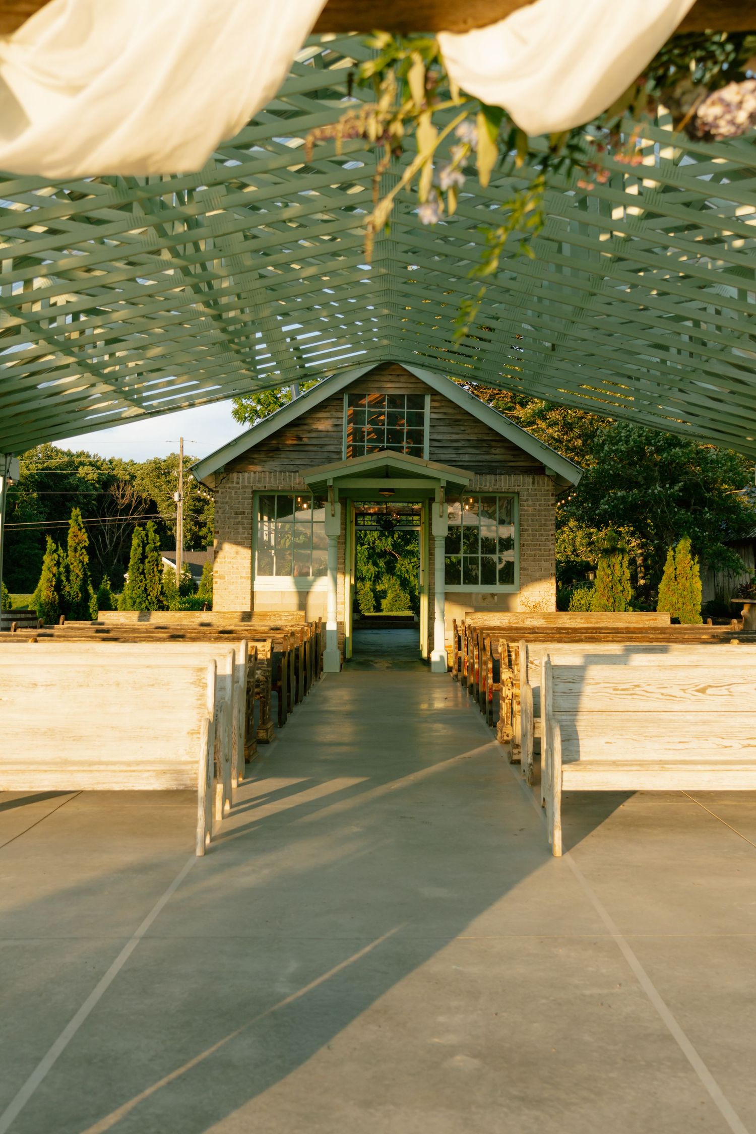 Elegant outdoor wedding aisle with wooden pews under lattice canopy leading to charming brick chapel.