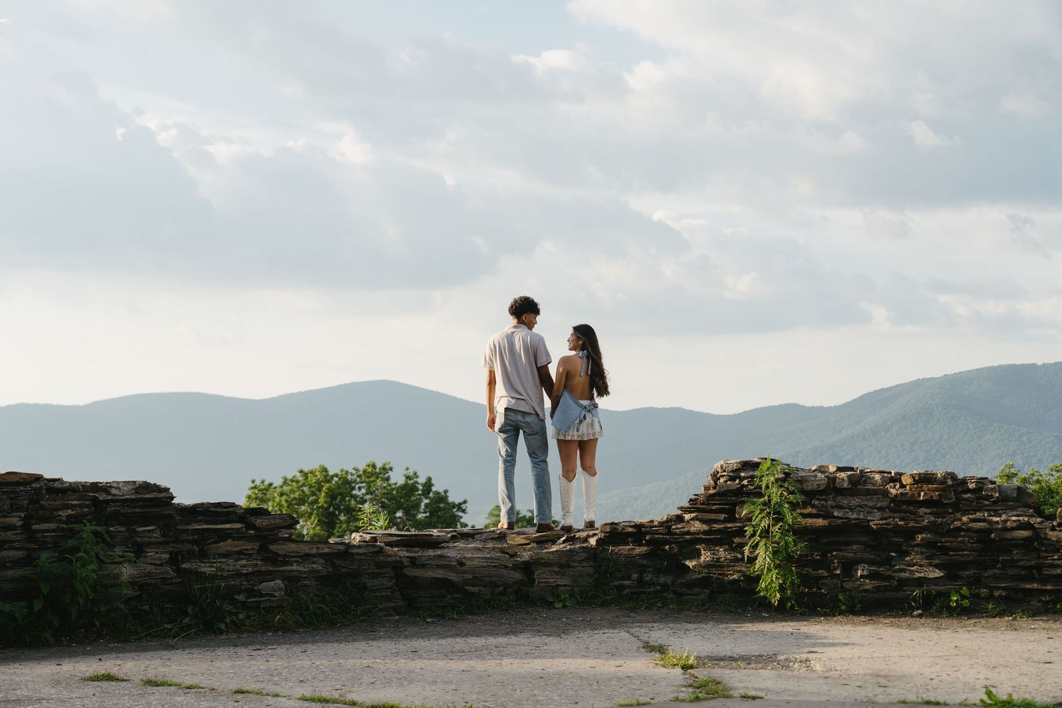 A couple stands hand-in-hand on a stone overlook gazing at a scenic mountain landscape.