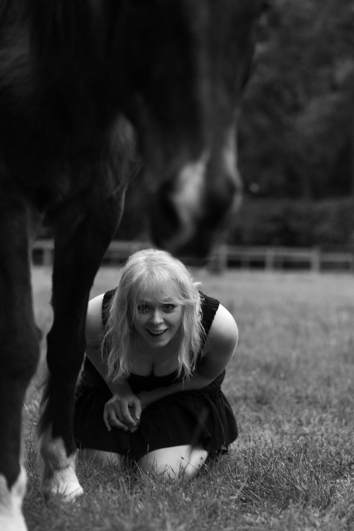 Black and white photo of smiling blonde woman crouching on grass near dark horse legs in outdoor paddock.
