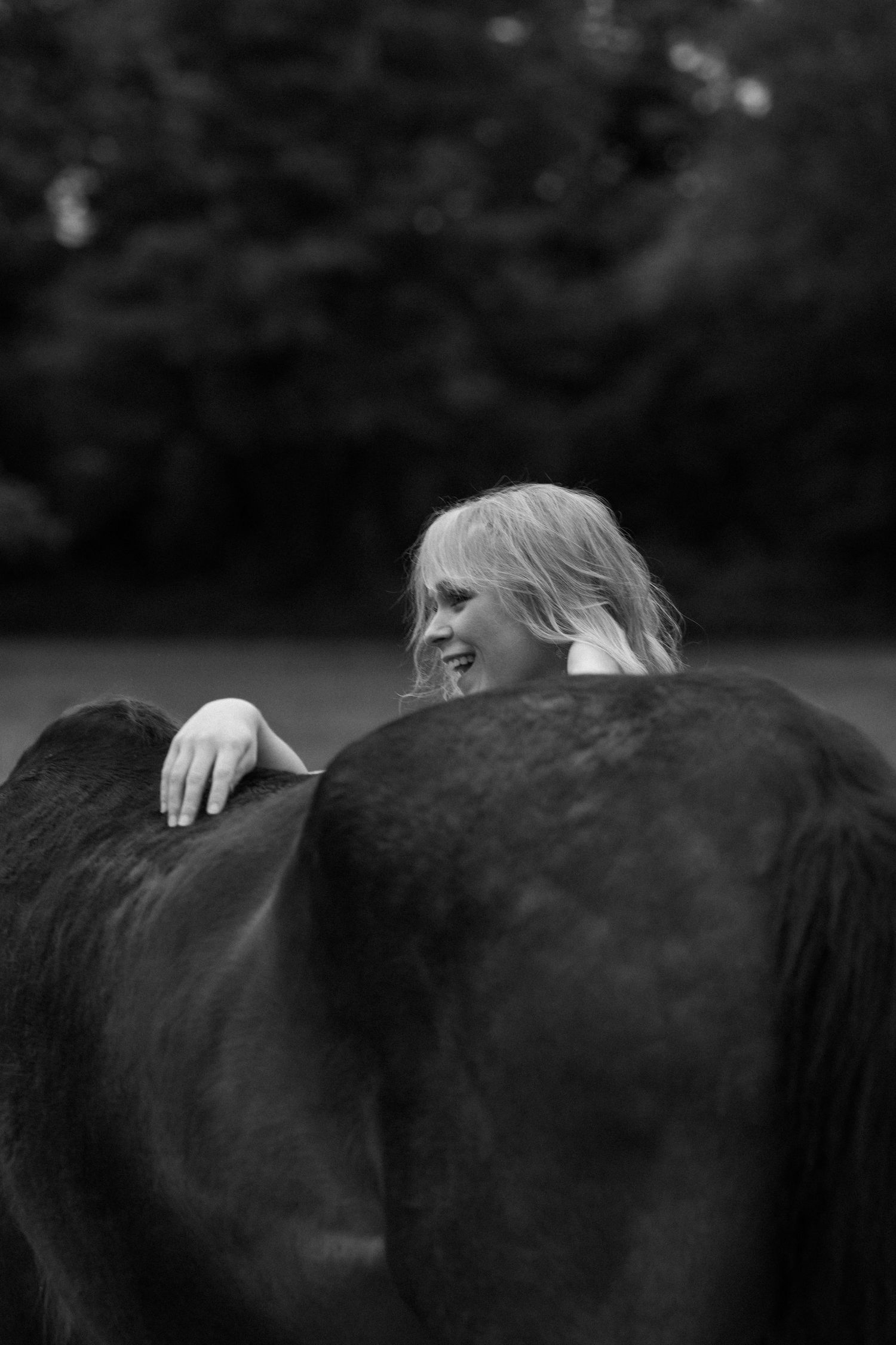 Black and white photo of blonde woman smiling behind dark horse outdoors.