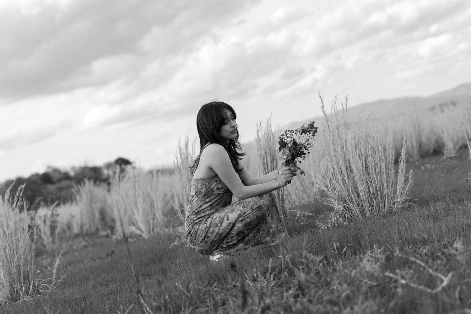 Black and white photo of a woman crouching in a field, holding wildflowers, with tall grass and cloudy sky behind her.