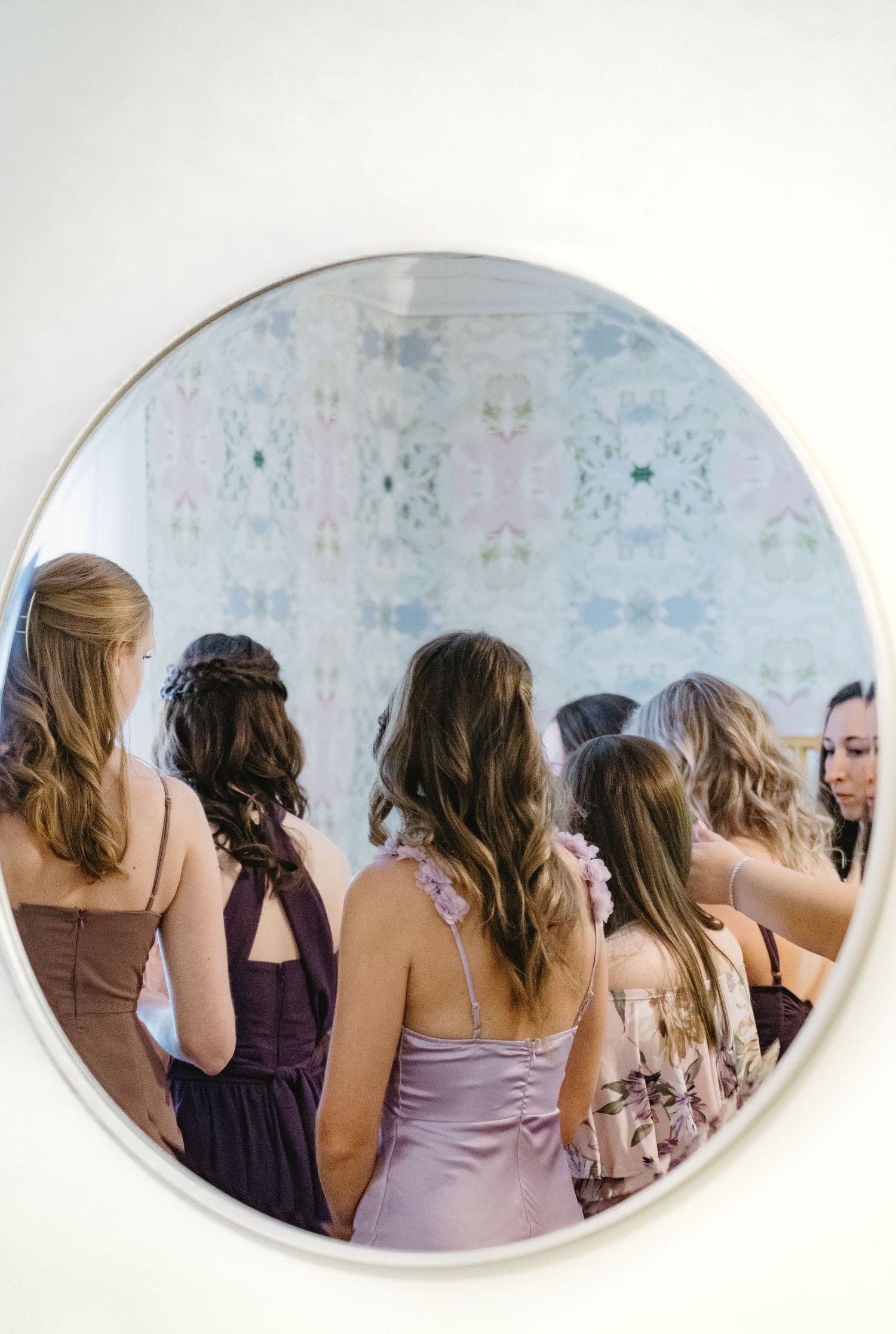 Group of bridesmaids in purple and mauve dresses viewed from behind reflected in large round wall mirror.