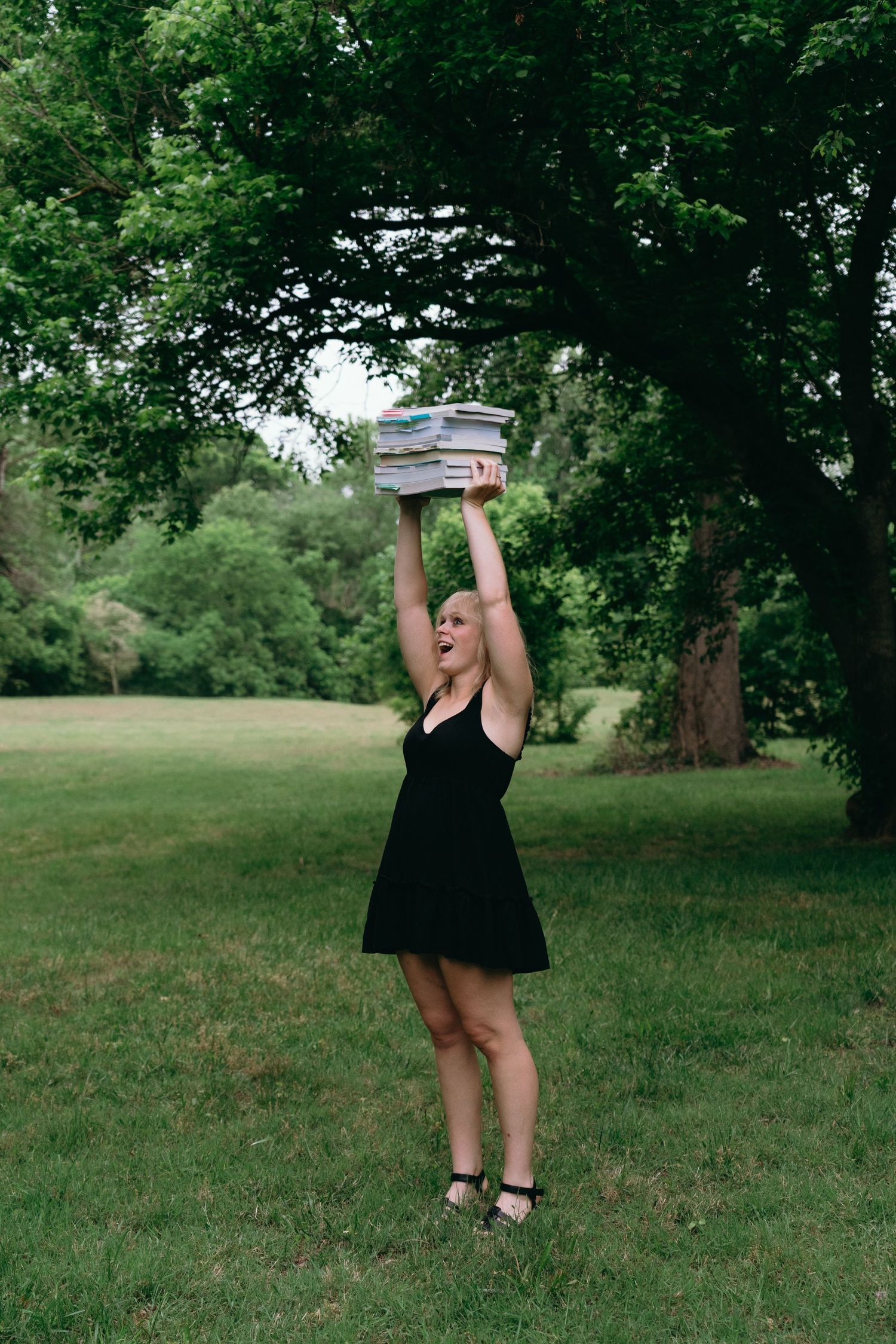 A joyful woman in a black dress raises a stack of books overhead in a lush green park.