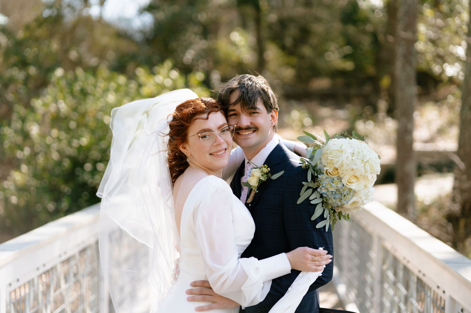 Smiling bride with red hair and flowing veil embraces groom holding white bouquet on white bridge.