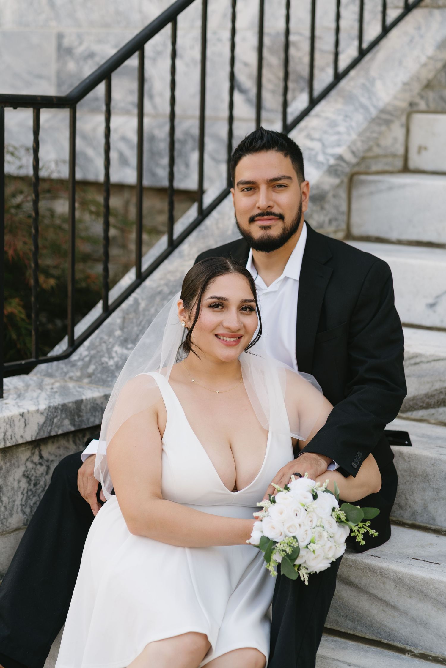 A smiling bride and groom pose on stone steps, bride holding a white bouquet of flowers.