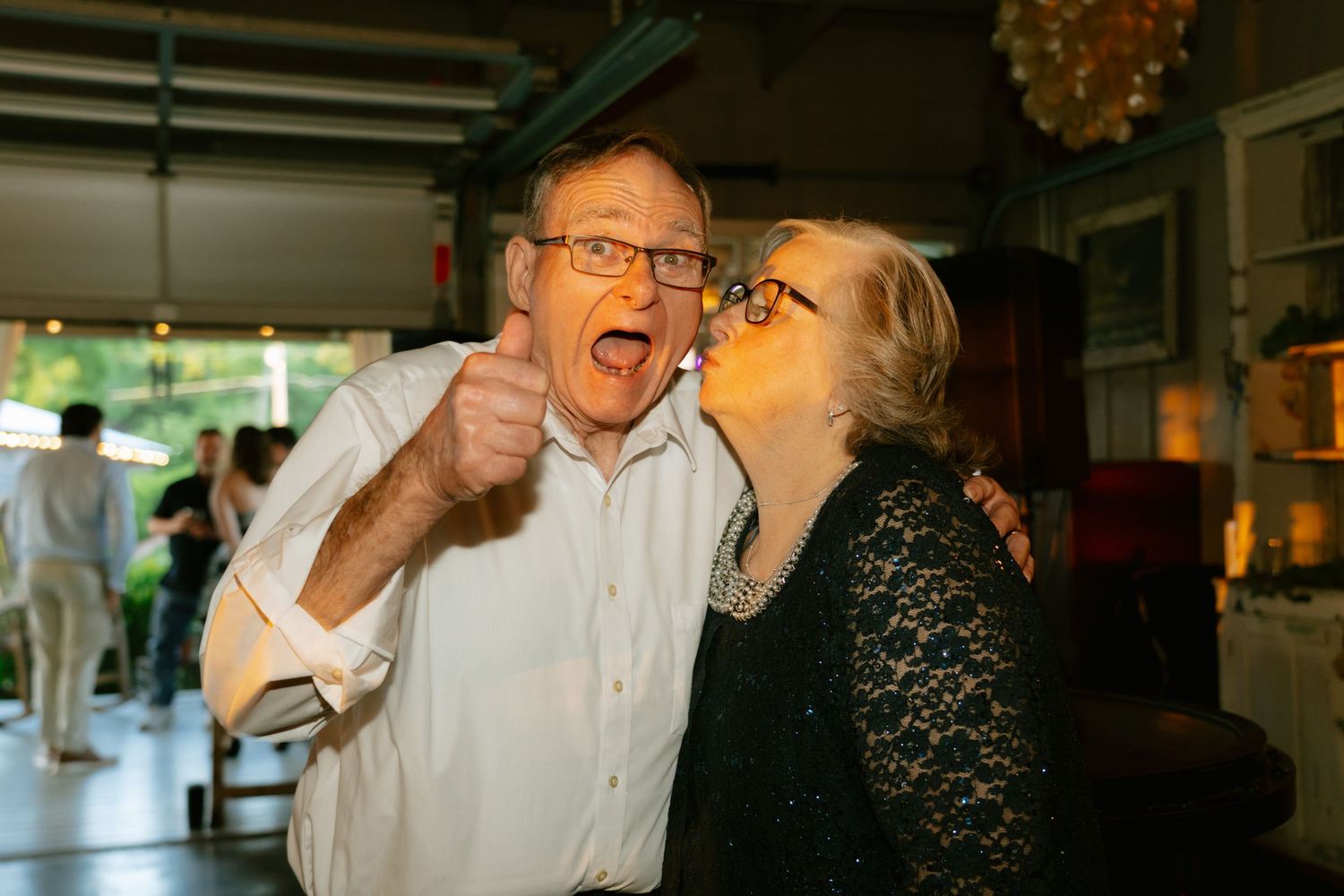 Happy couple celebrates at a party, man gives thumbs up while woman kisses his cheek indoors.