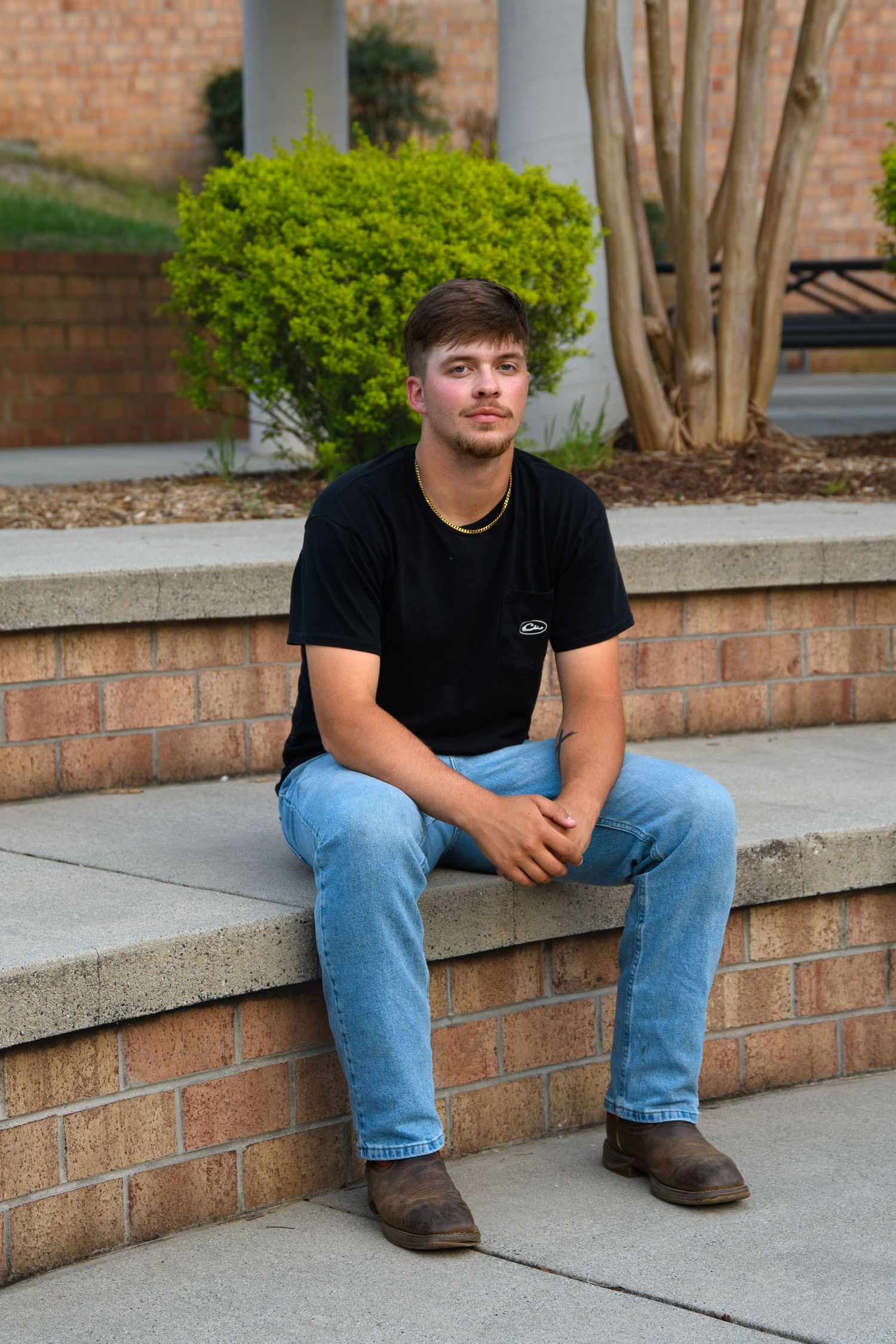 Young man in black t-shirt and jeans sits on brick steps outdoors near green shrubs.