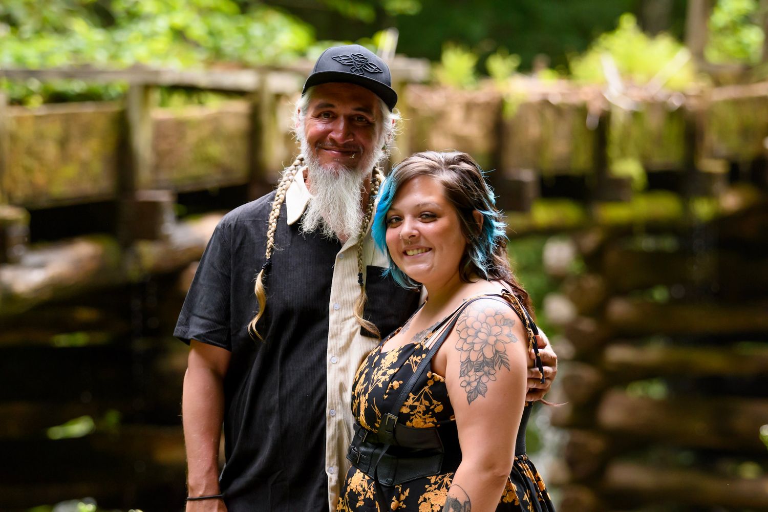 Smiling couple posing outdoors near a wooden bridge surrounded by lush green trees in a forest setting.