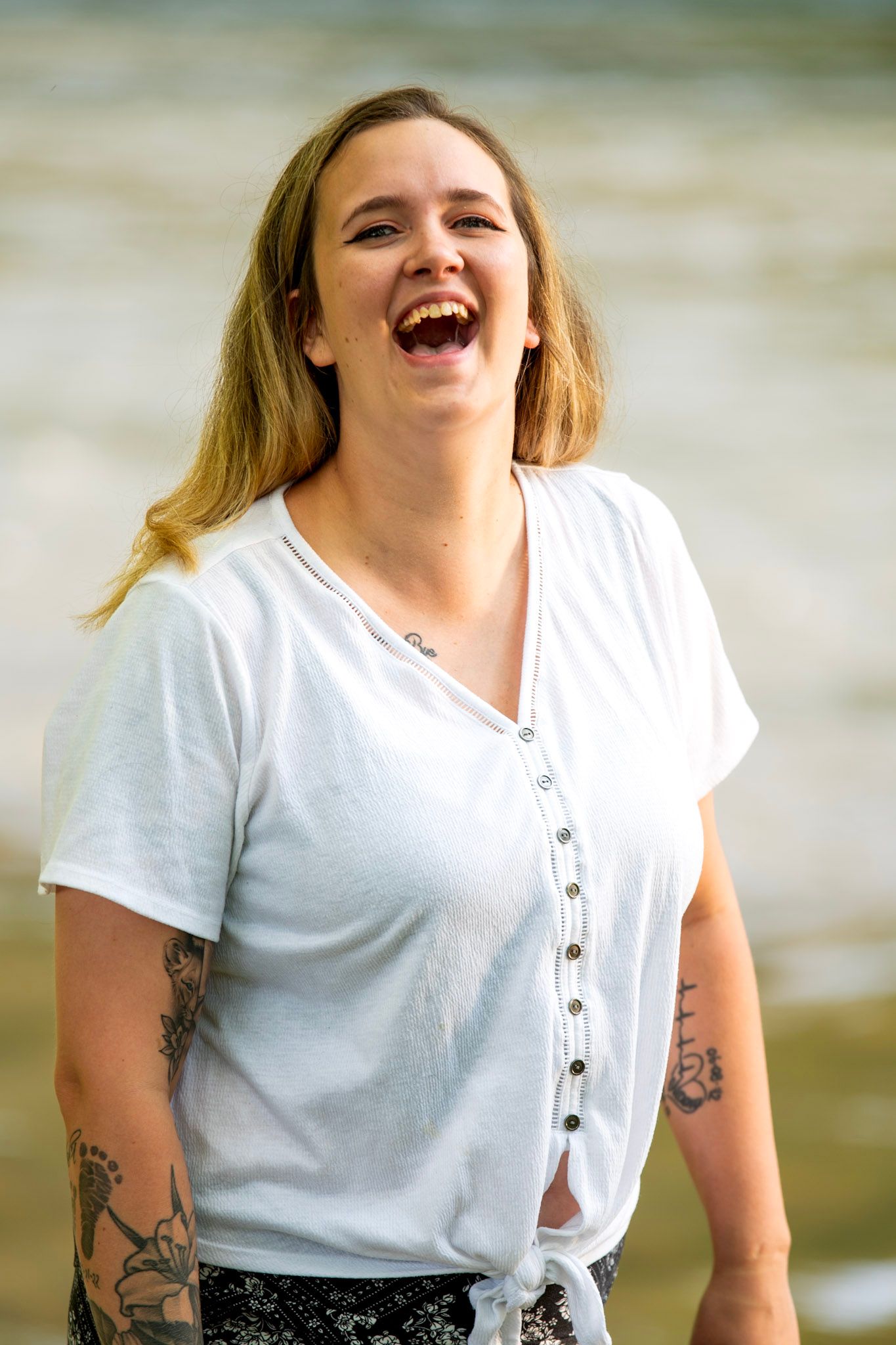 Laughing young woman with tattoos wearing a white t-shirt standing near a body of water outdoors.