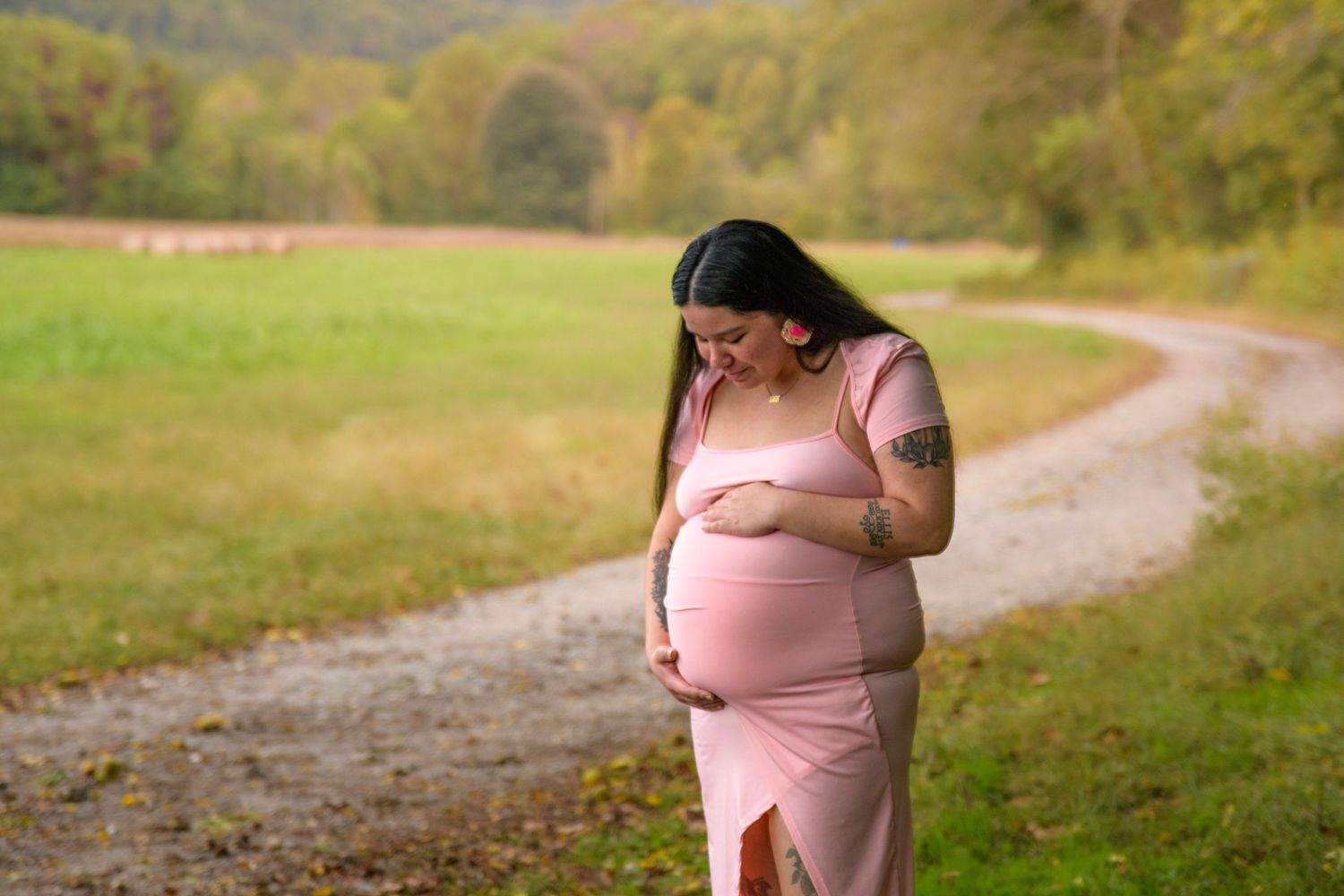 Pregnant woman in pink dress cradles baby bump on a scenic rural gravel path.