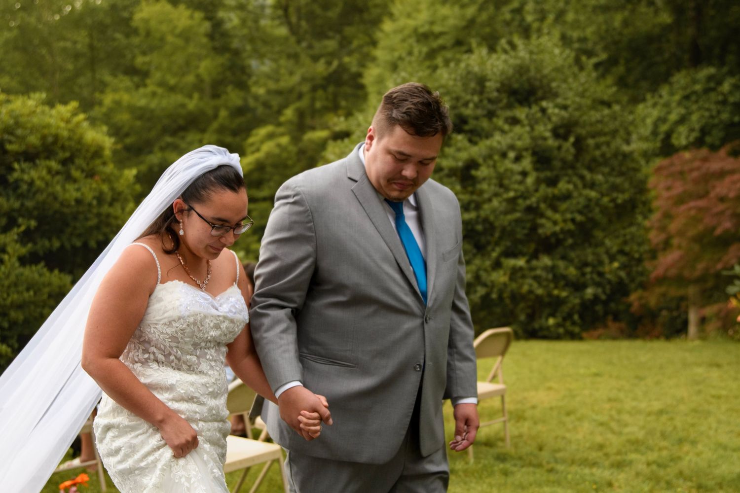 Bride in white gown and veil holds hands with groom in gray suit during outdoor wedding ceremony.