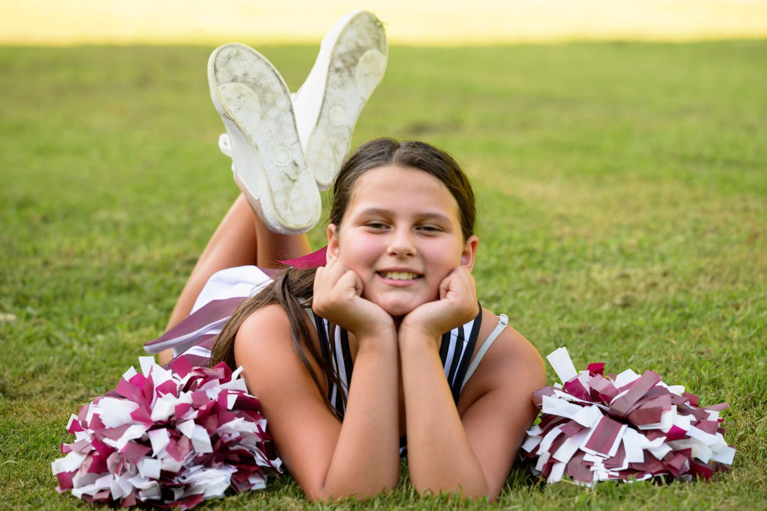 Young cheerleader in uniform lying on grass field, smiling with pom-poms, wearing white cheer shoes.