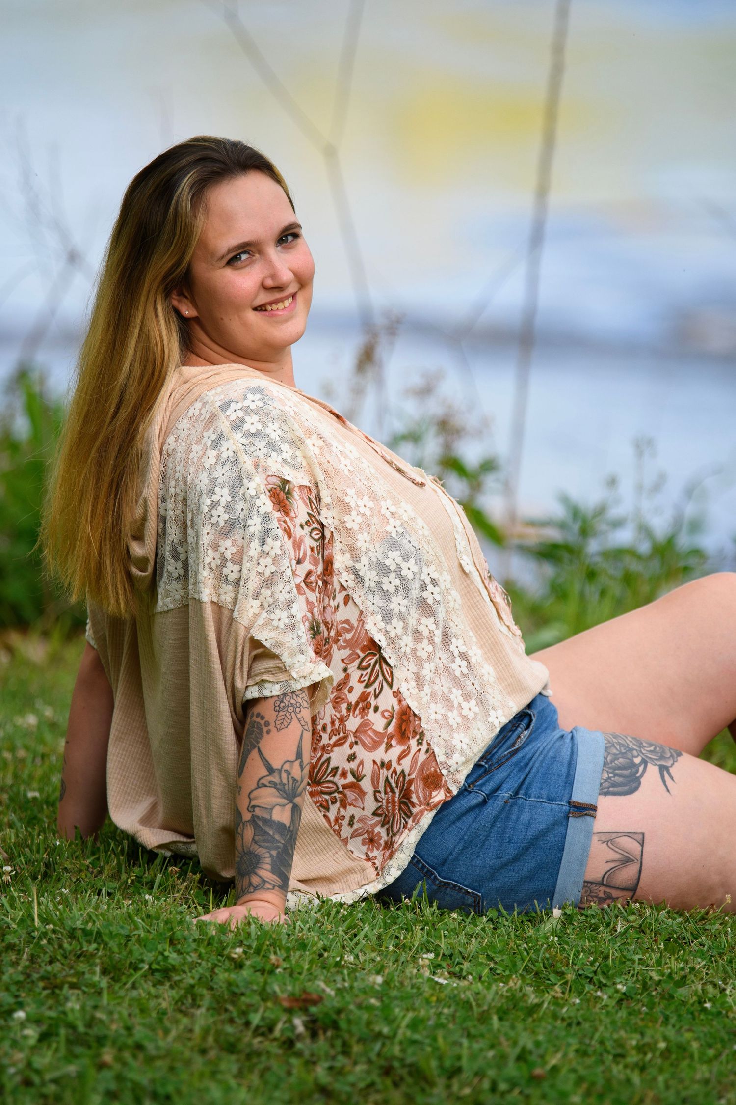 Tattooed young woman with long blonde hair sitting on grass in denim shorts and lace top, smiling.