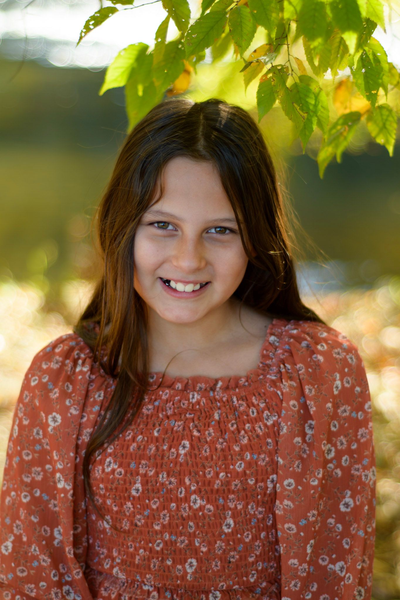 Smiling brunette girl in floral top standing under green leaves outdoors.