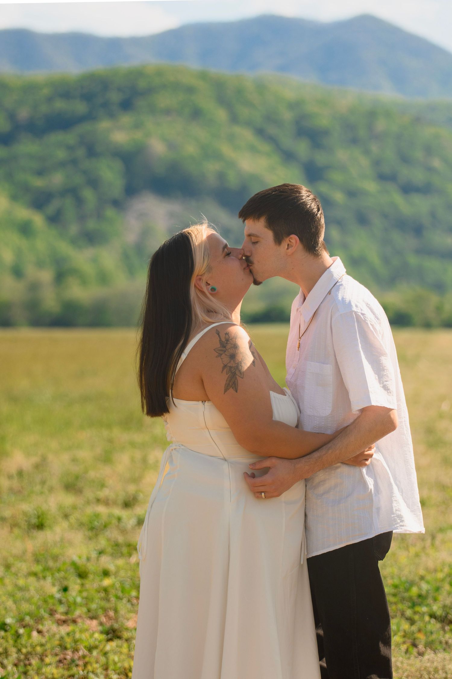 Couple sharing a romantic kiss in an open field with lush green mountains in the background.