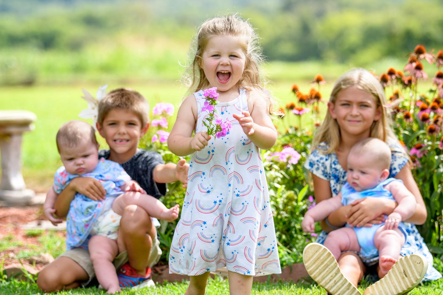 Five children pose outdoors among pink flowers, with two holding babies and a girl in a rainbow dress laughing.