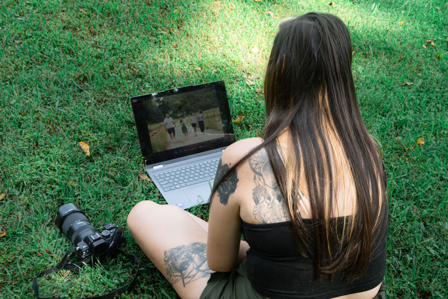 Tattooed woman sitting on grass in a park, viewing photos on a laptop with a camera beside her.
