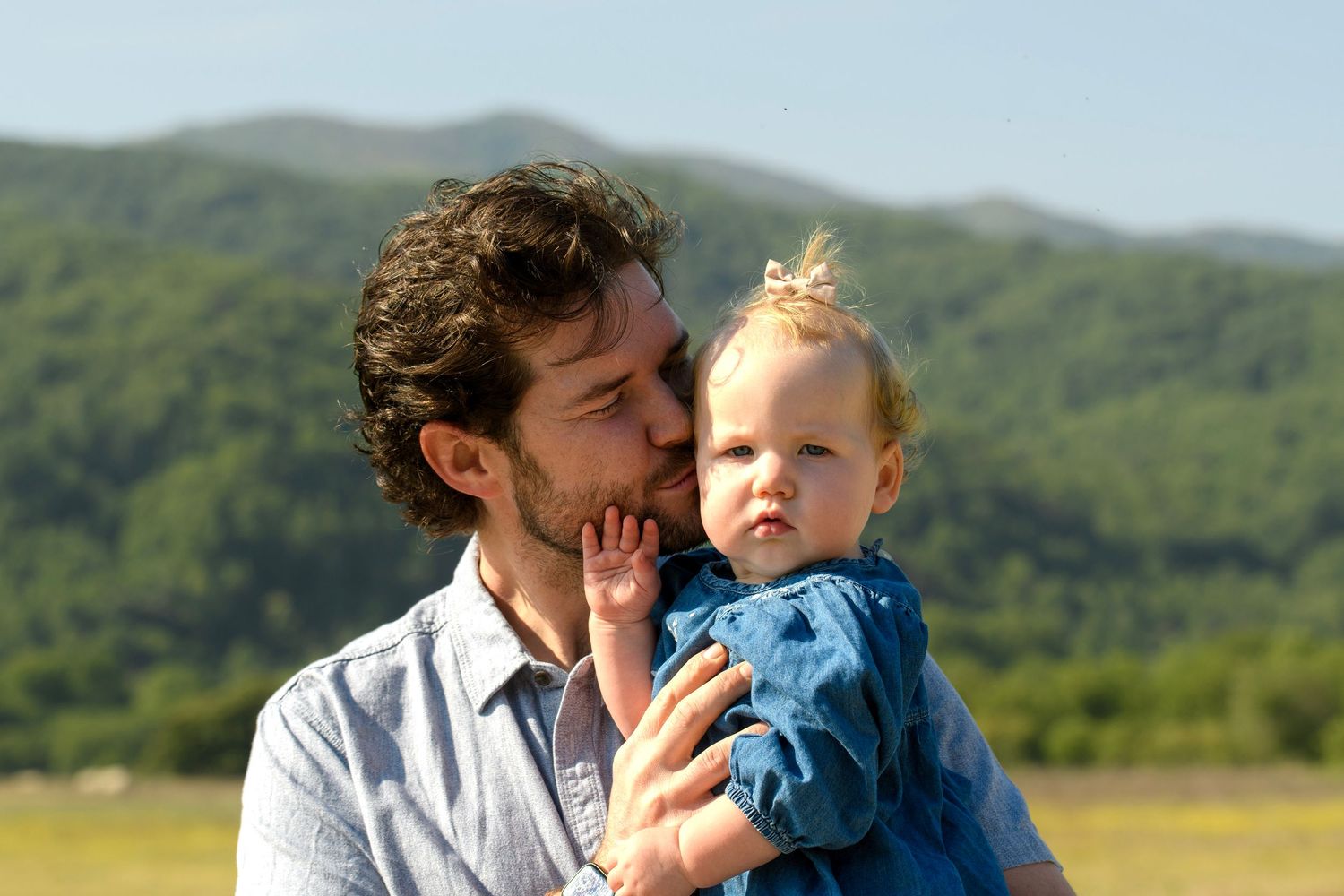 A man with curly hair kisses a baby girl outdoors with green mountains in the background.
