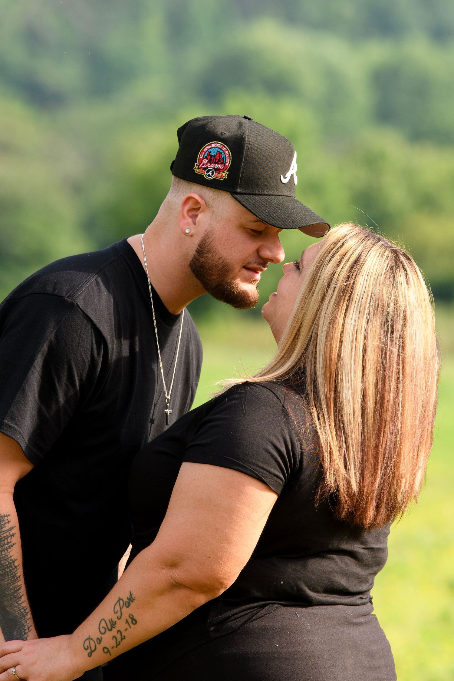 Couple in matching black outfits sharing a romantic moment outdoors, man wearing an Alabama baseball cap with green hills behind.