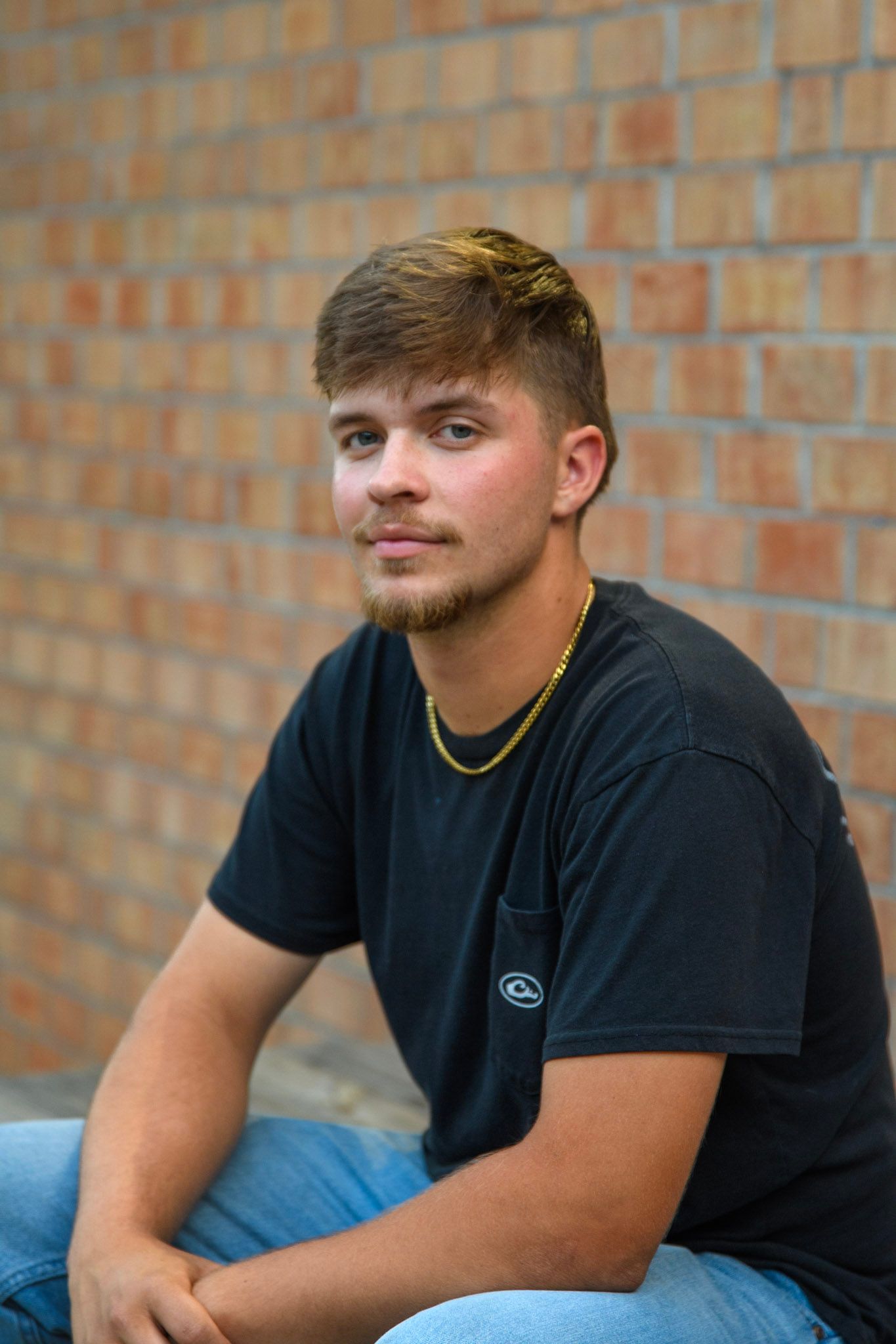 Young man in black t-shirt and gold chain sits against a brick wall, posing for a casual outdoor portrait.