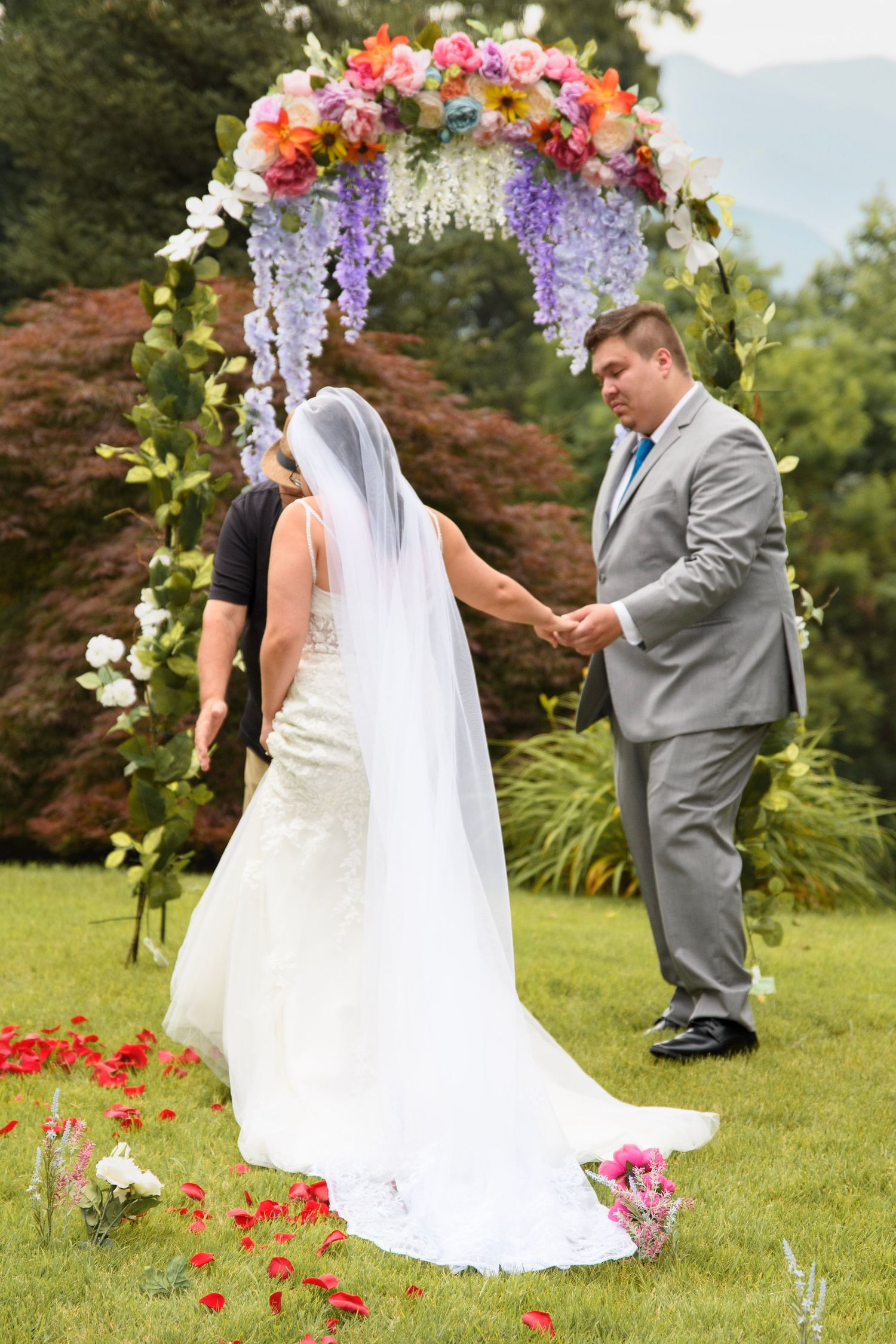 Bride and groom holding hands at outdoor wedding ceremony under colorful floral arch with wisteria on green lawn.