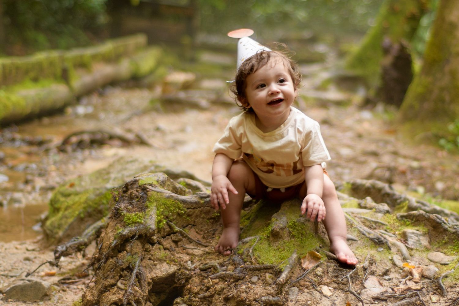 A happy toddler in a party hat sits barefoot on mossy rocks in a lush forest setting.