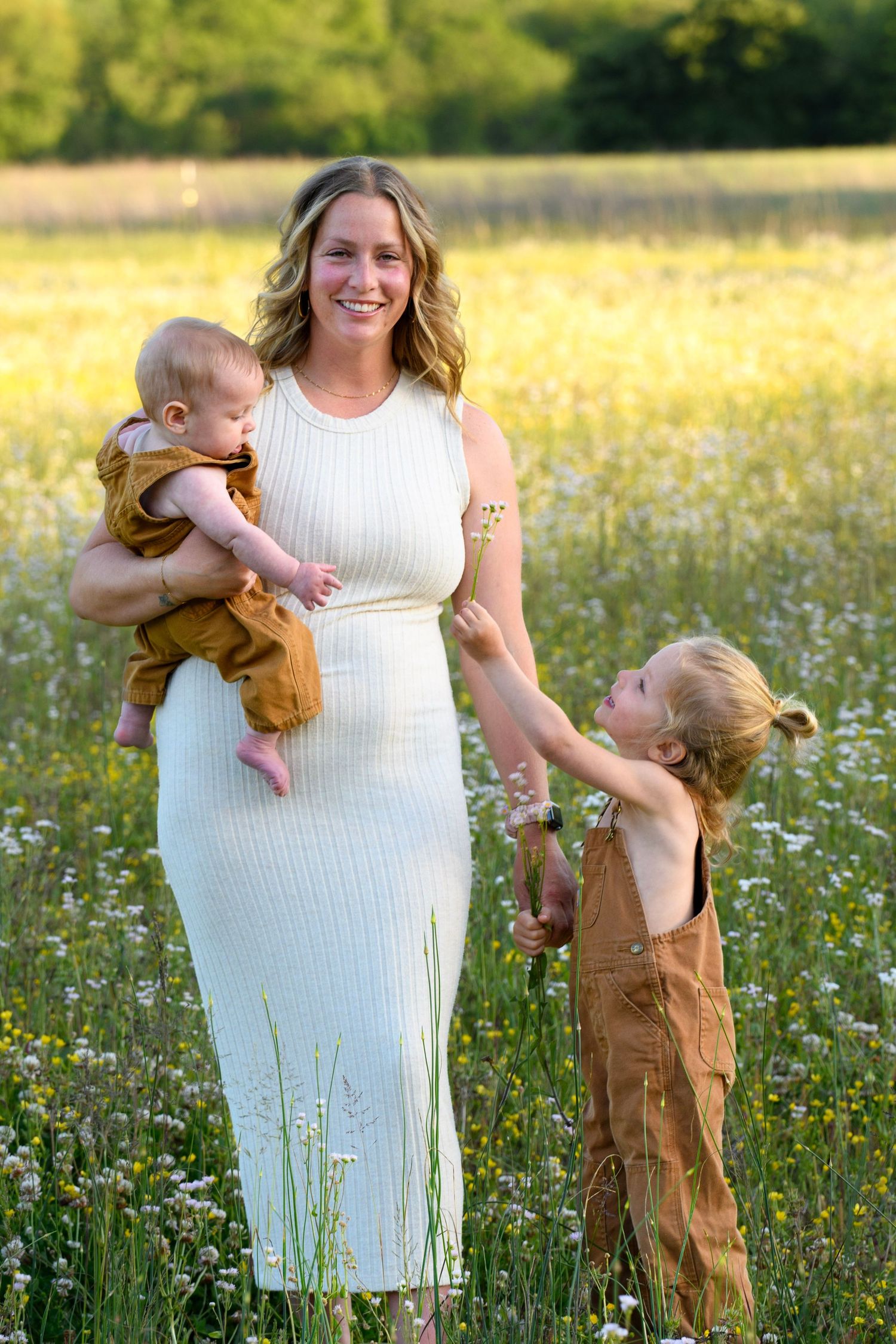 Smiling mother in white dress holding baby while toddler reaches up in wildflower field.