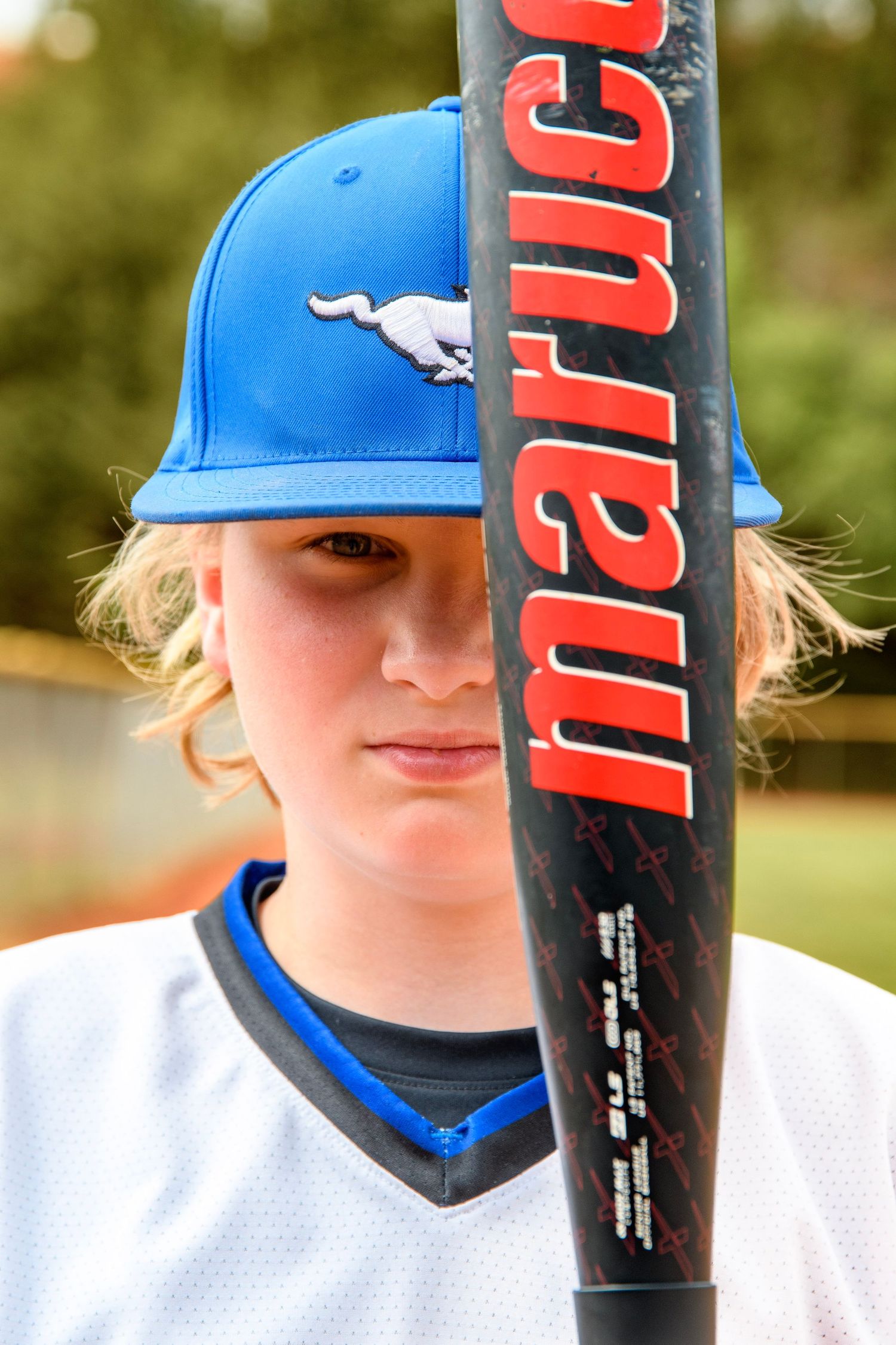 Young male baseball player holding a Marucci bat in front of his face wearing a blue Mustangs cap.