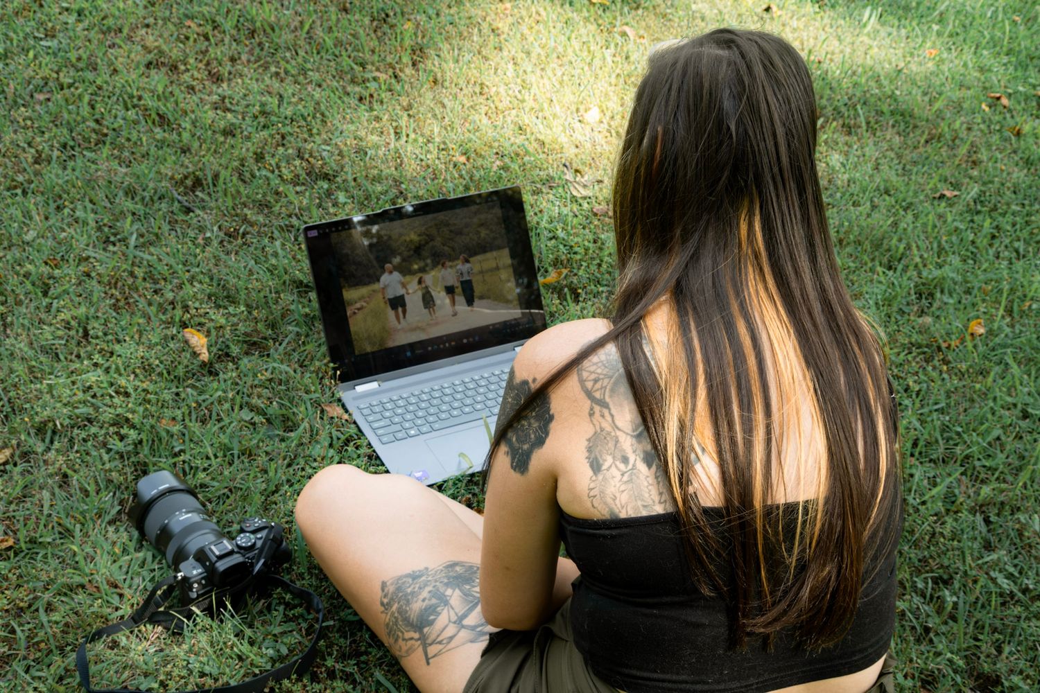 Tattooed woman sitting on grass in a park, viewing photos on a laptop with a camera beside her.