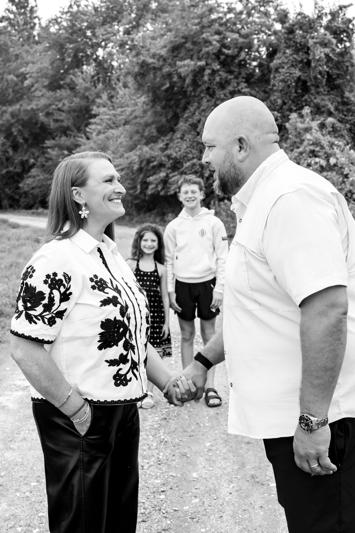 Black and white photo of a couple holding hands and smiling, with two children standing in the background outdoors.