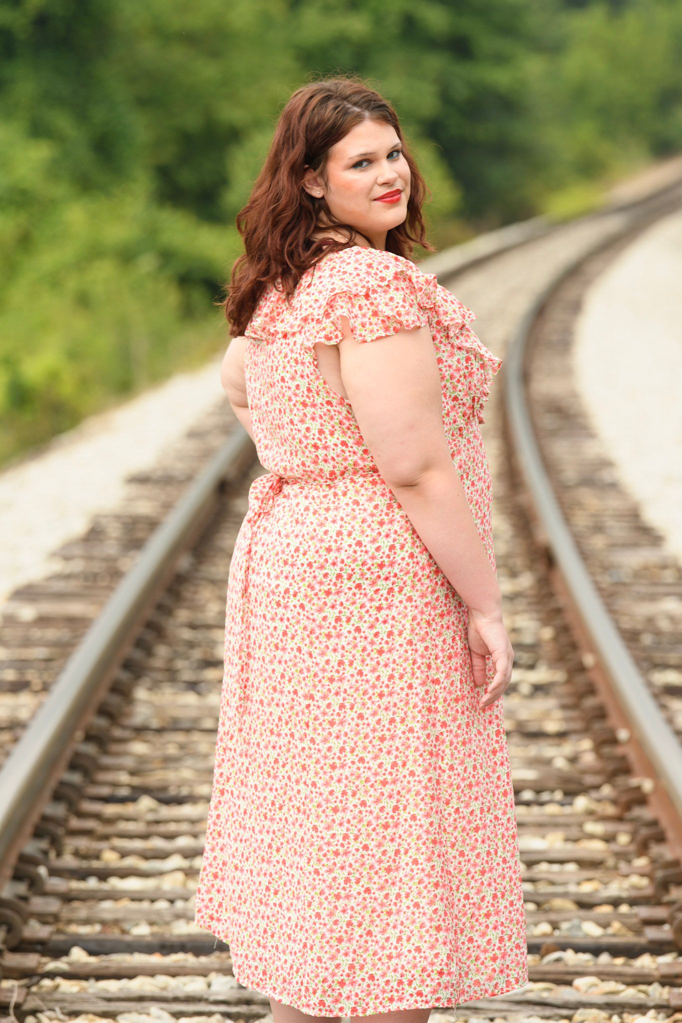 Plus-size woman in floral pink dress posing on railroad tracks surrounded by lush green trees.